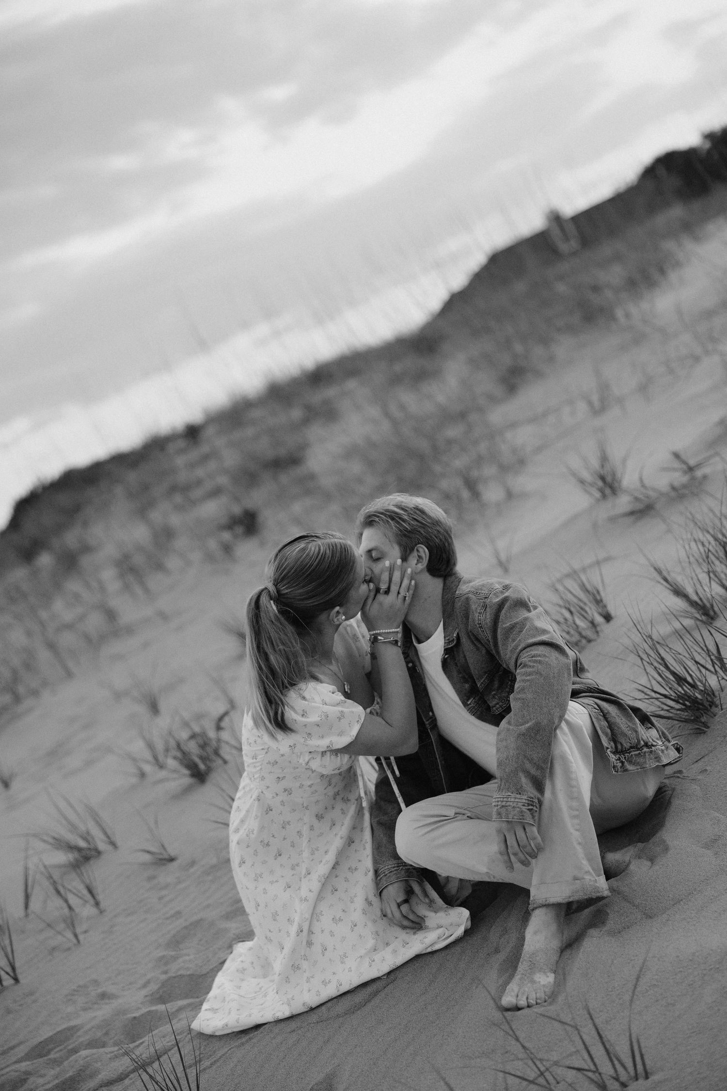 Couple shares intimate kiss sitting on sandy beach dunes in black and white portrait.