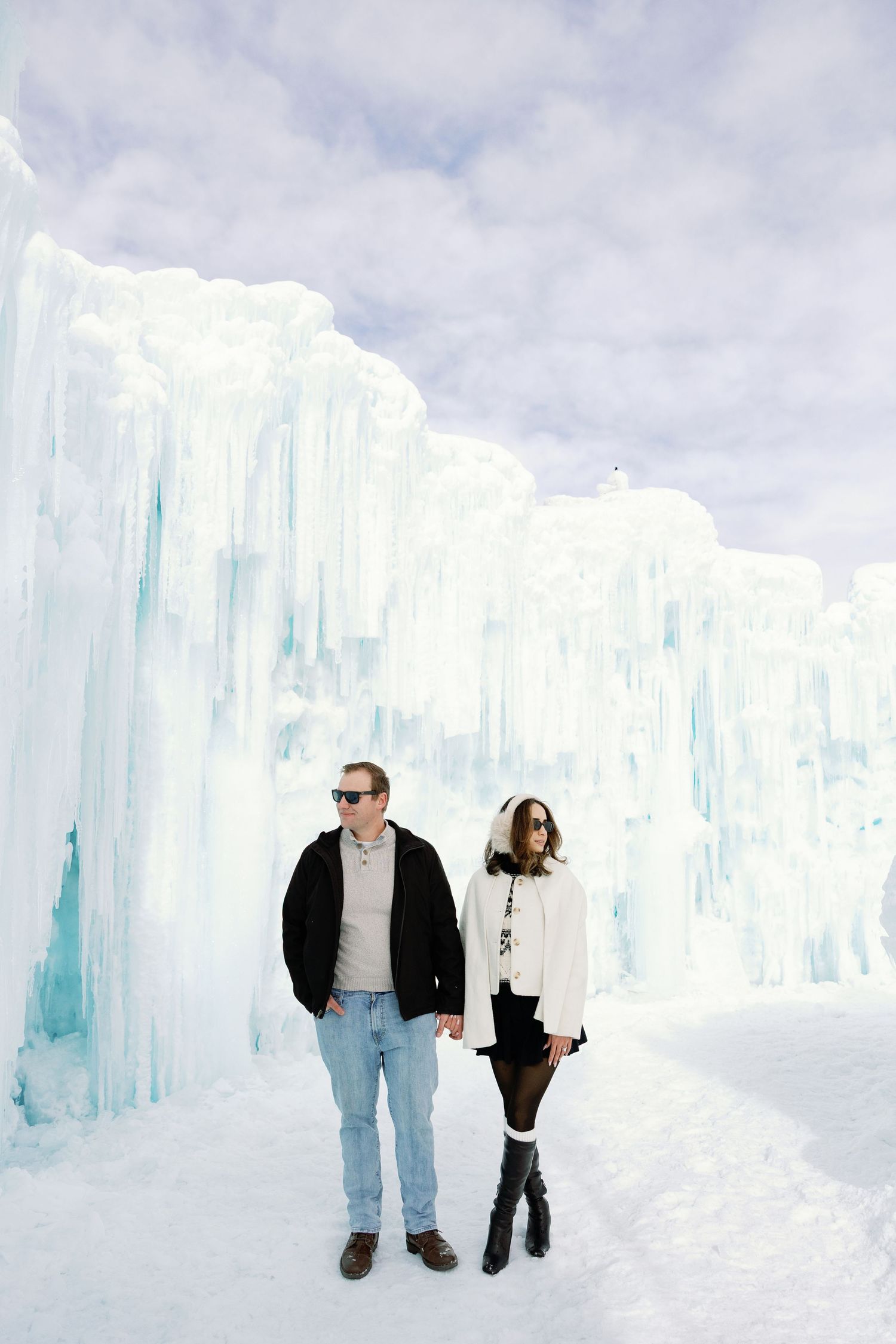 Stylish couple walking hand in hand along snowy path beside towering ice castle walls.