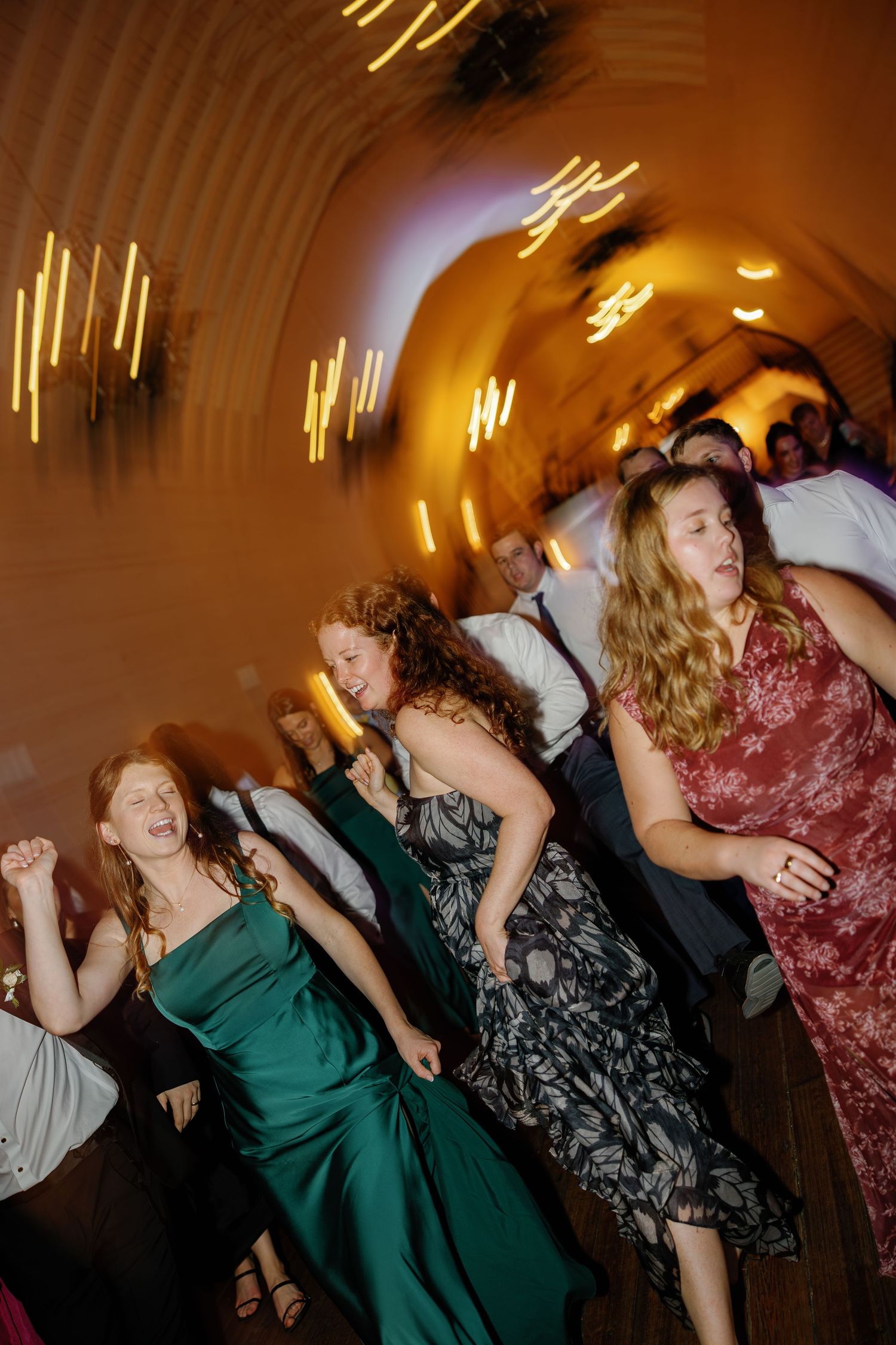Wedding guests in formal attire dance energetically on the dance floor during reception celebration.
