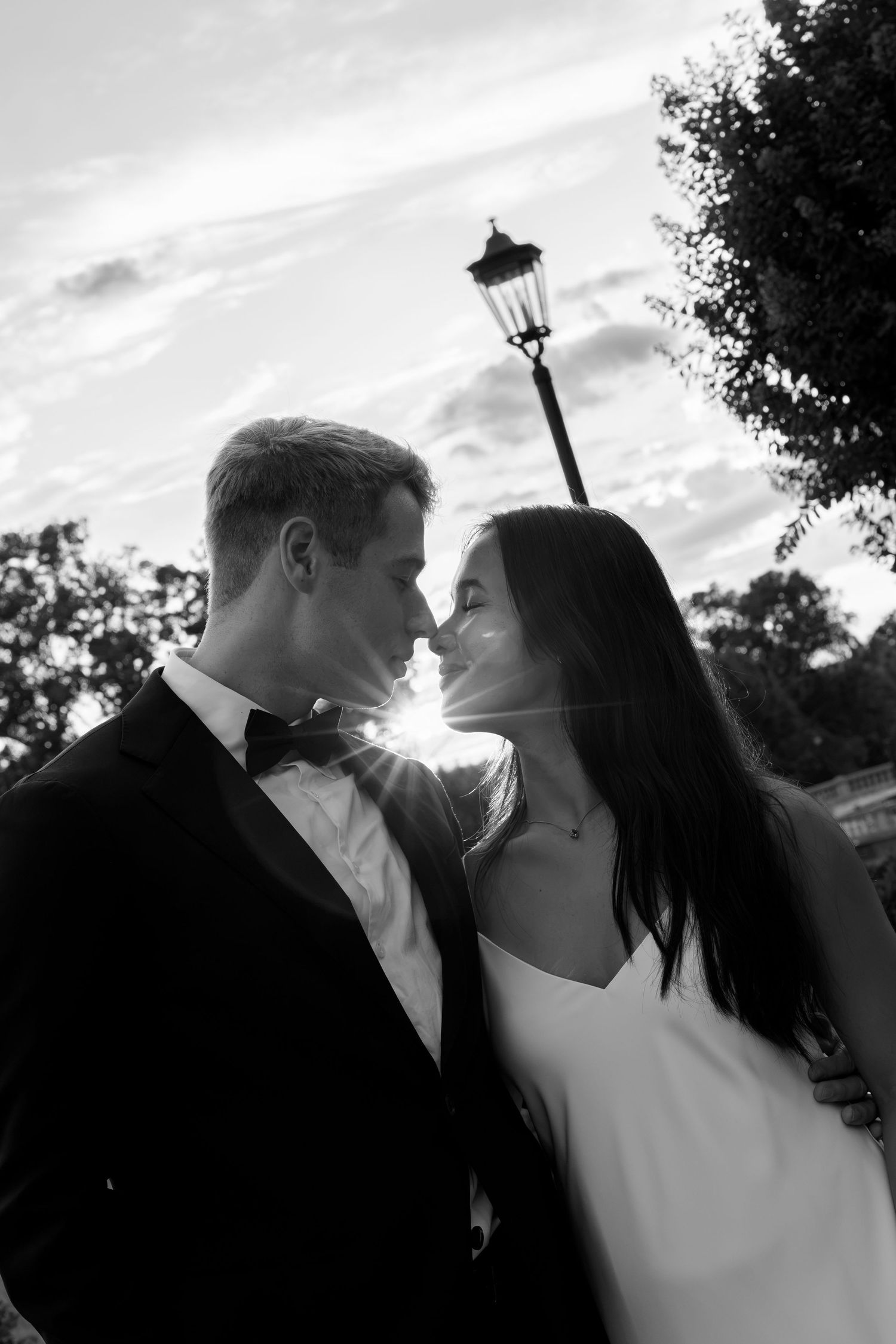 Black and white photo of bride and groom almost kissing with sunburst between them at sunset.