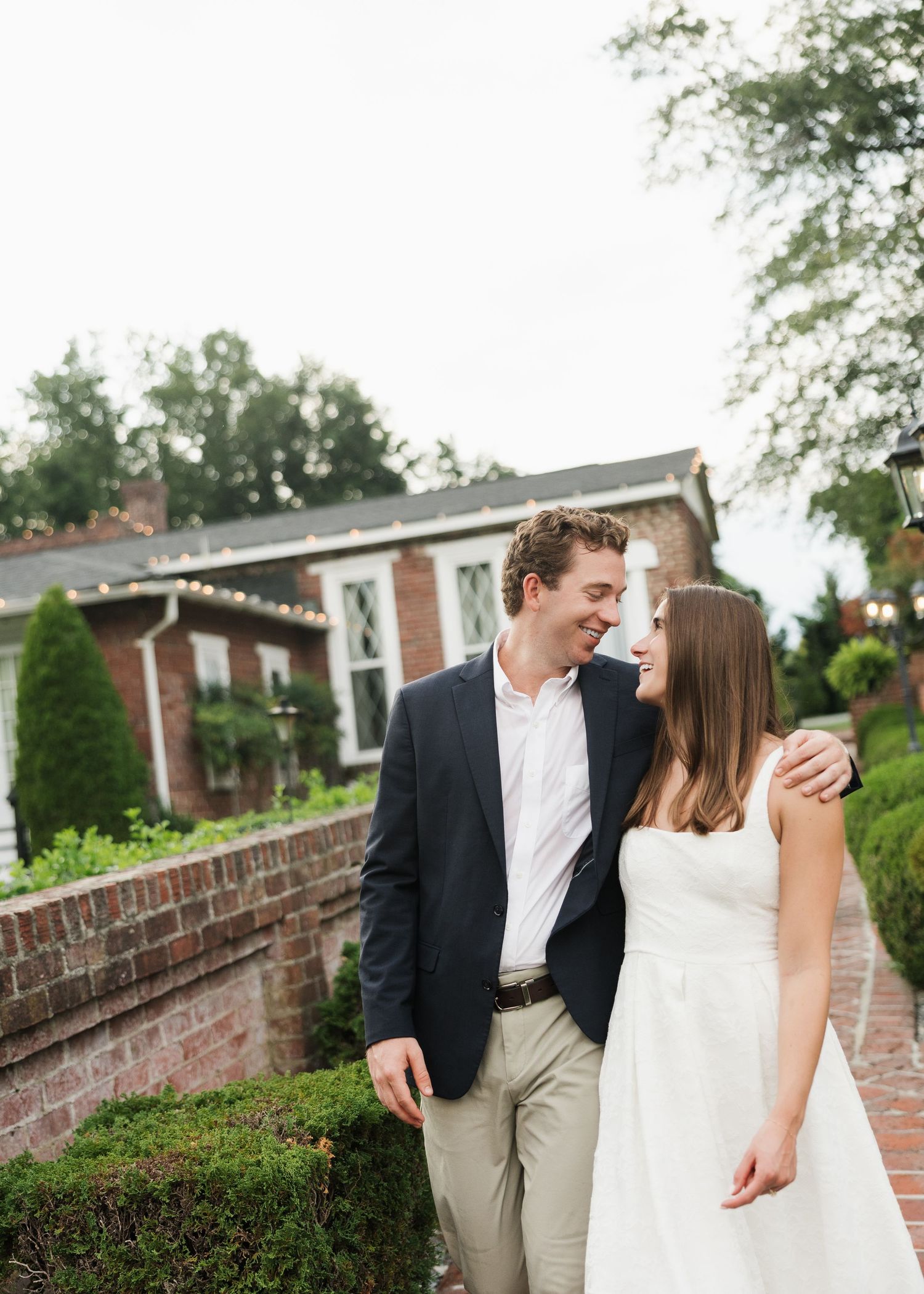 A smiling couple in elegant attire walks together near a brick building with string lights and lush greenery.