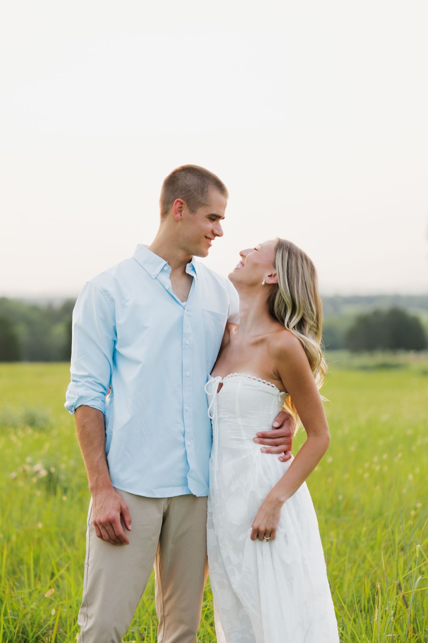 Romantic couple gazing at each other in a lush green meadow, man in light blue shirt, woman in white strapless dress.