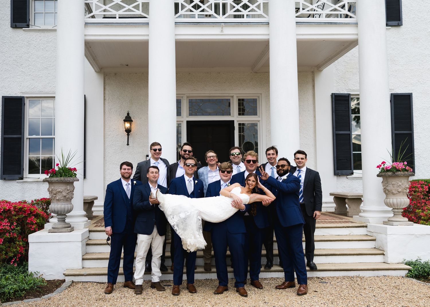 Groomsmen in navy suits carry bride on steps of white colonial mansion at outdoor wedding celebration.
