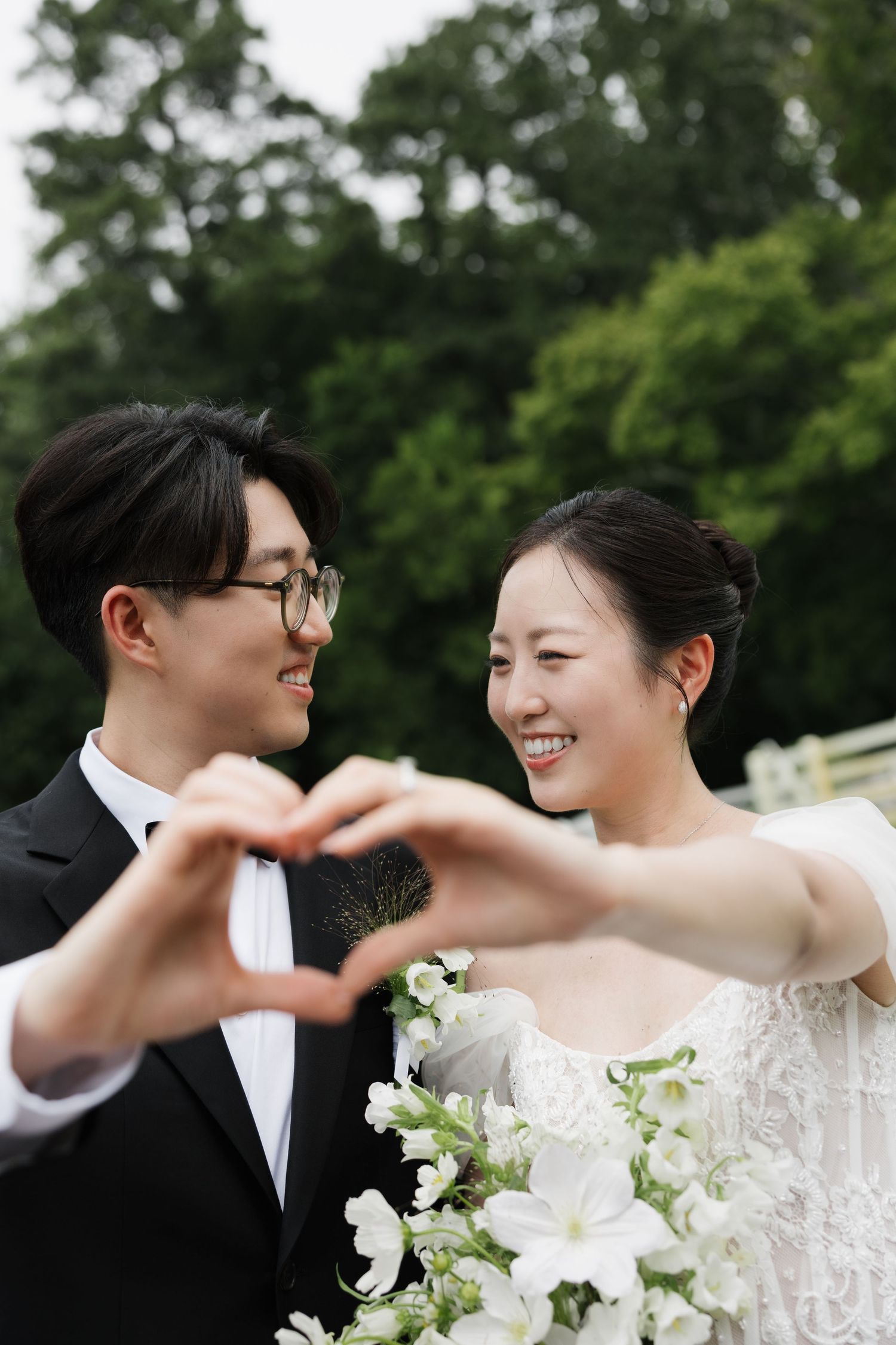 A smiling Asian bride and groom in wedding attire make a heart shape with their hands outdoors.