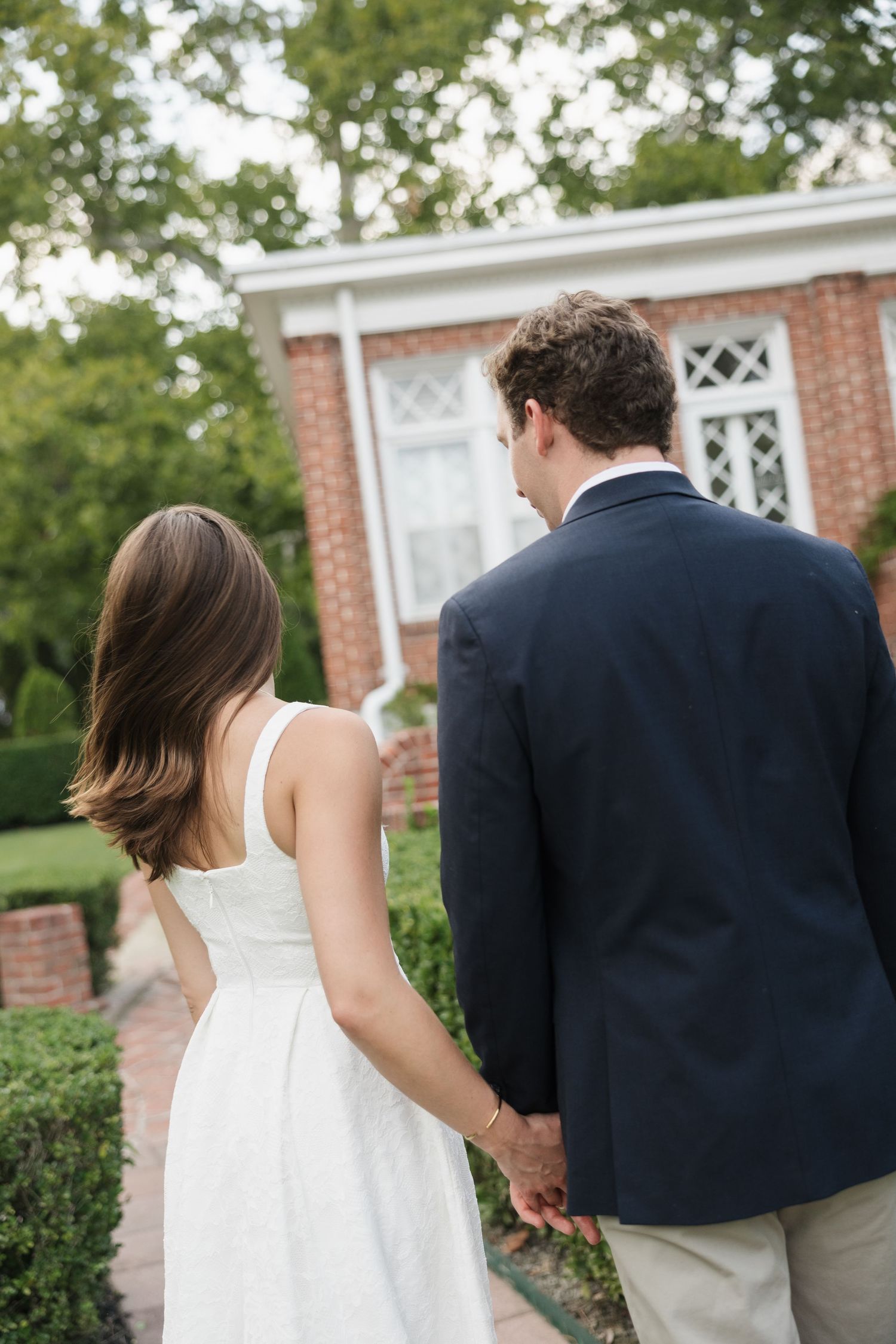 Couple holding hands walking toward brick building, woman in white dress and man in navy suit, rear view.