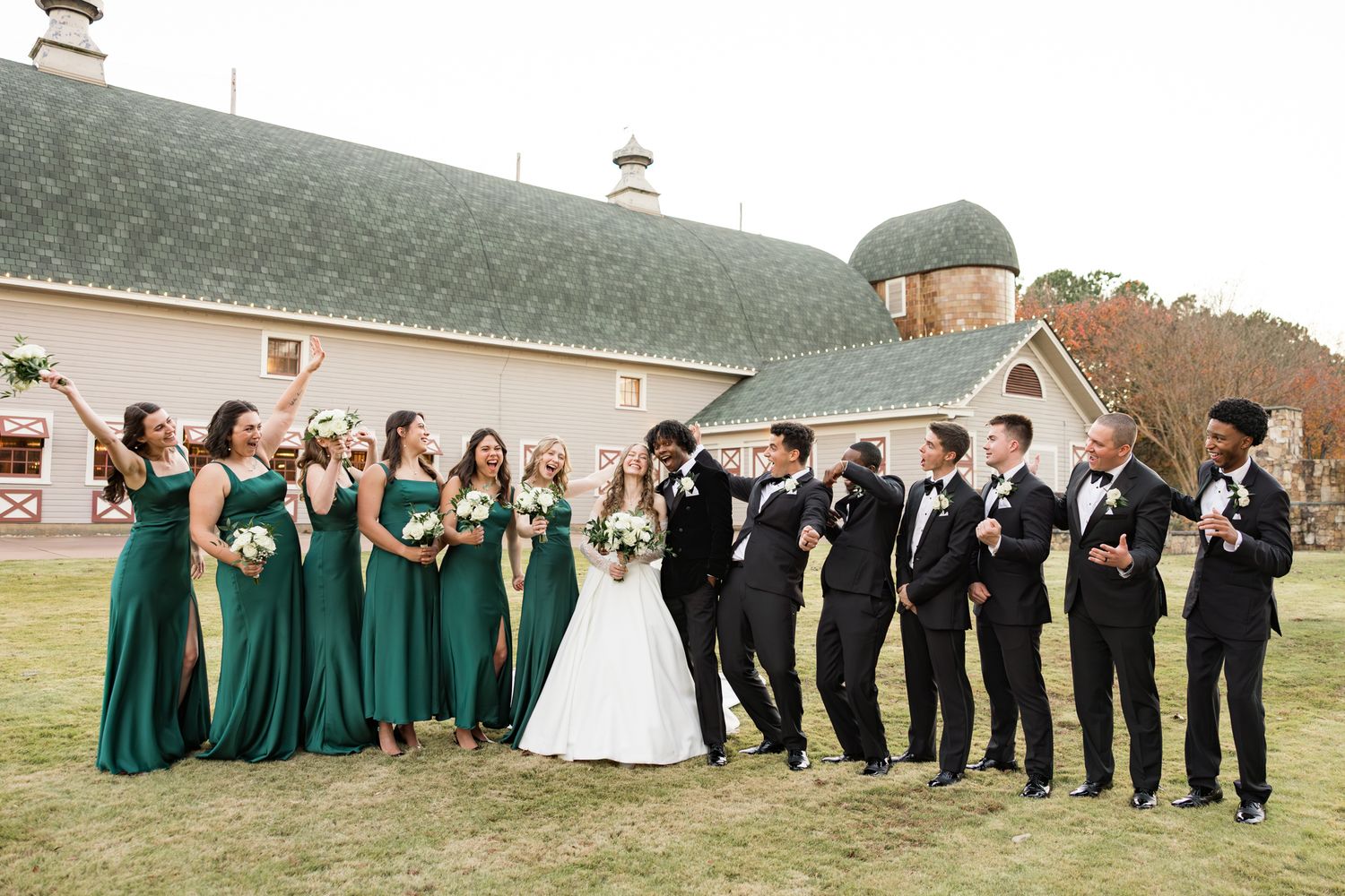 Wedding party in emerald green gowns and black tuxedos pose outside rustic barn venue.
