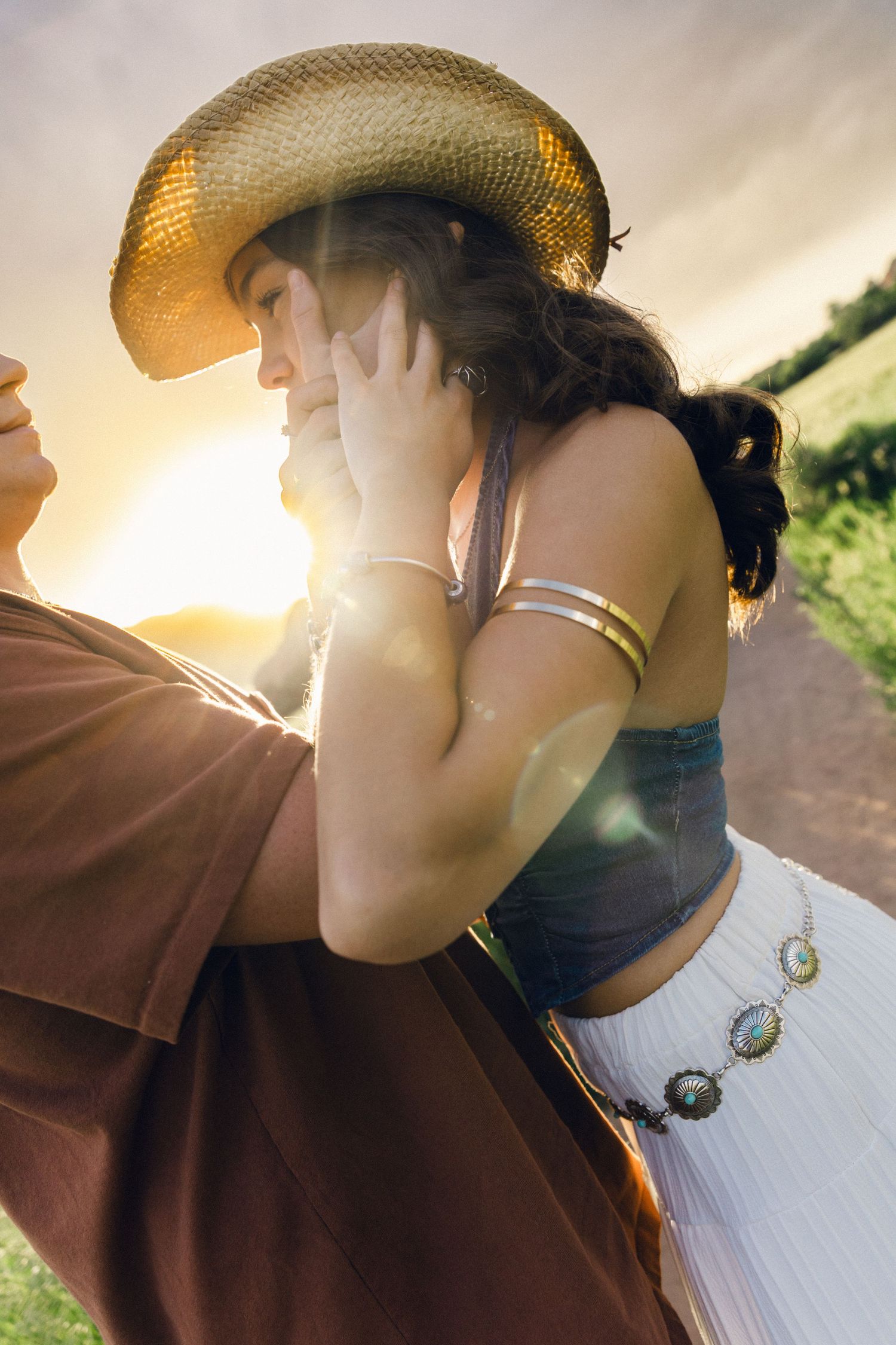 Woman in straw cowboy hat and crop top embraced by partner at golden sunset in rural countryside.