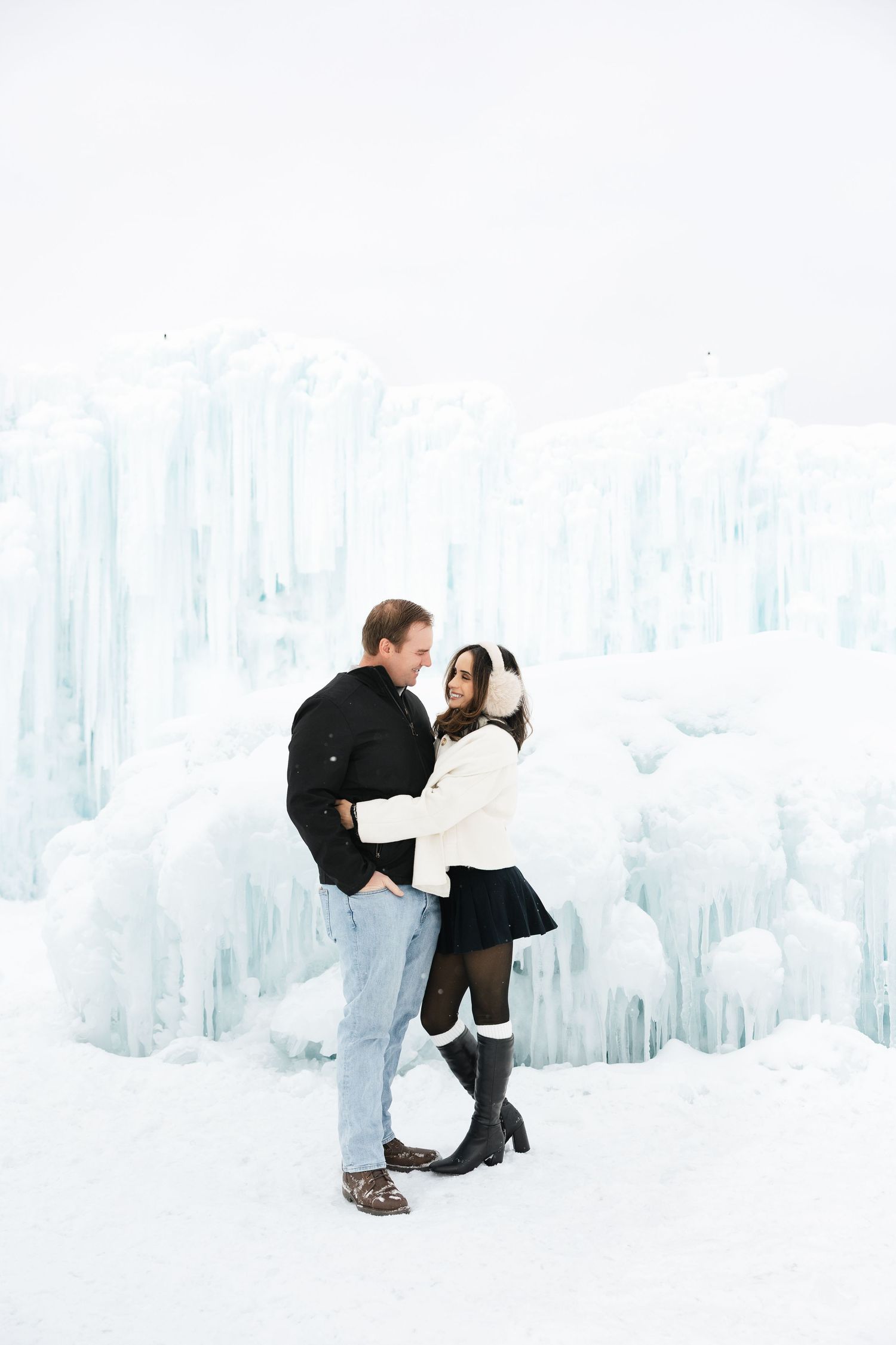 Romantic couple embracing and laughing in front of towering ice castle formations in winter.