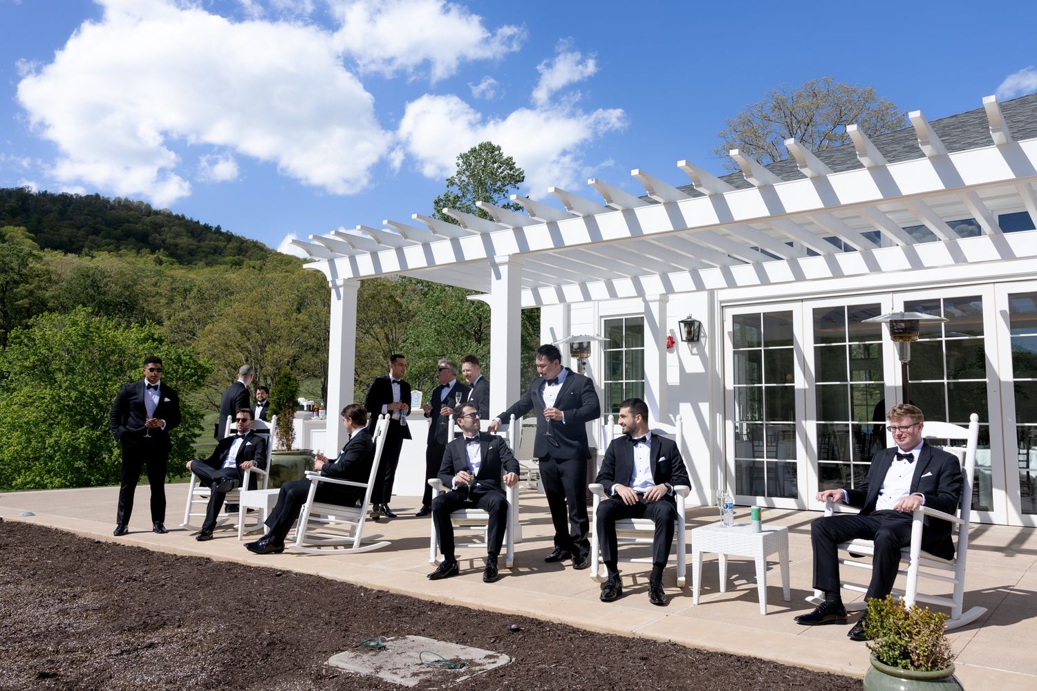 Groomsmen in tuxedos relaxing near a white pergola outdoor wedding venue with lush green hills and blue sky.