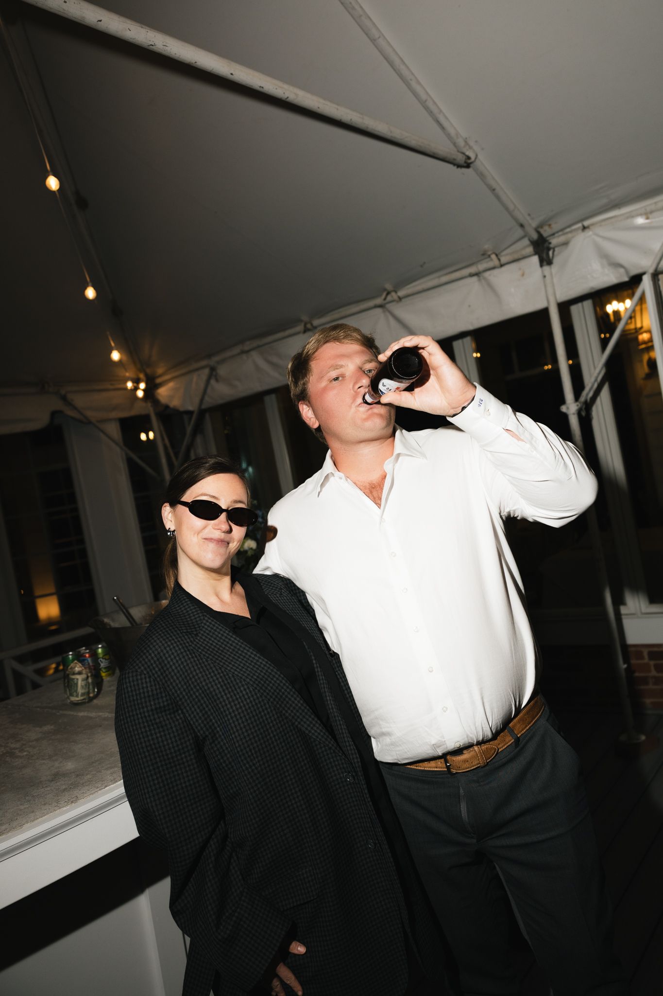 A woman in black sunglasses stands beside a man in a white shirt drinking from a bottle at an outdoor evening rehearsal dinner.