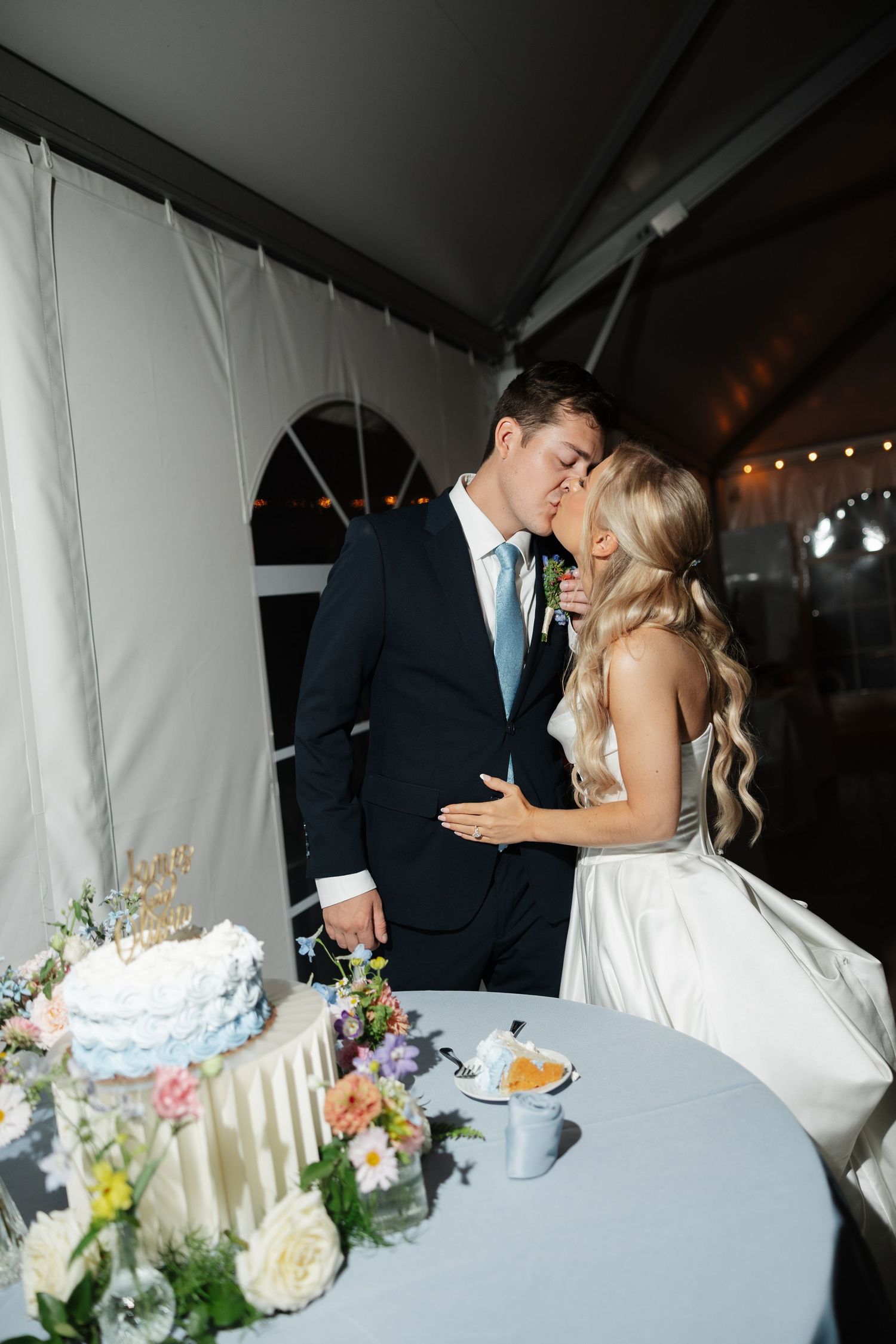 Bride and groom sharing a kiss while cutting their wedding cake under a tent reception venue.