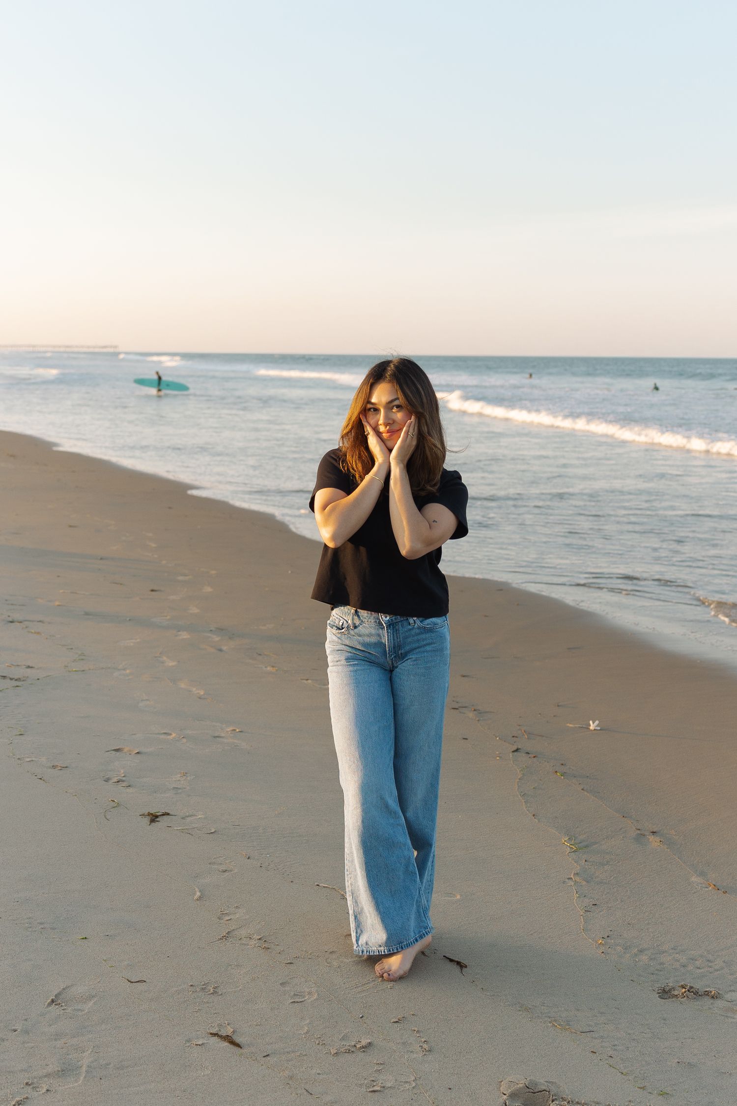 Young woman in black top and wide-leg jeans stands barefoot on sandy beach at sunset with ocean waves behind her.
