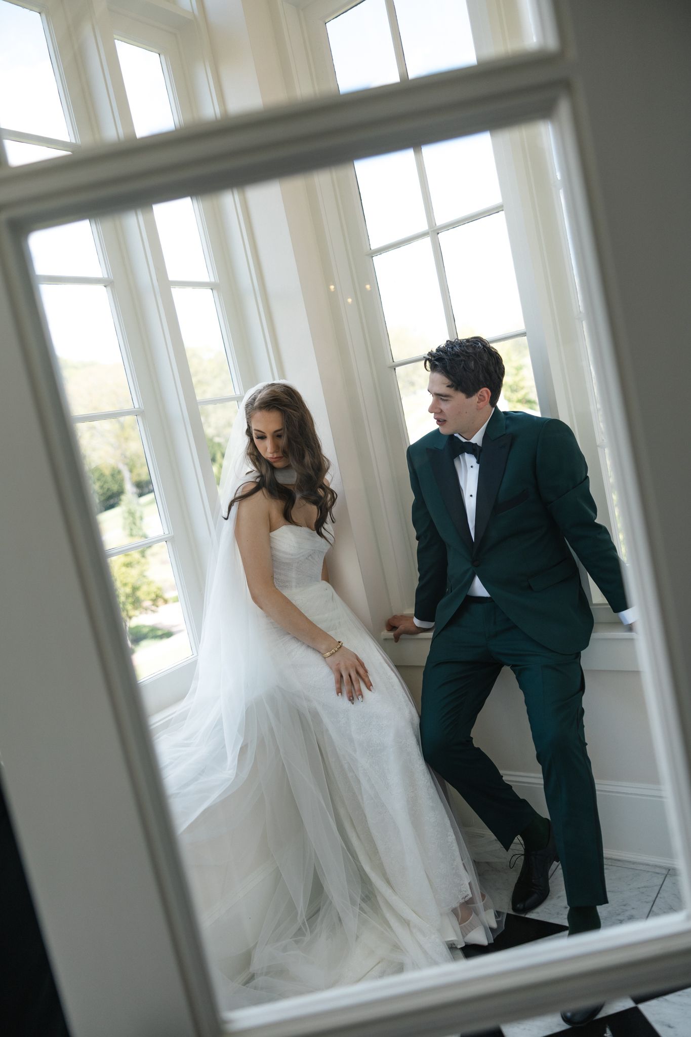 Bride in white gown and groom in green tuxedo reflected in mirror by sunlit windows.