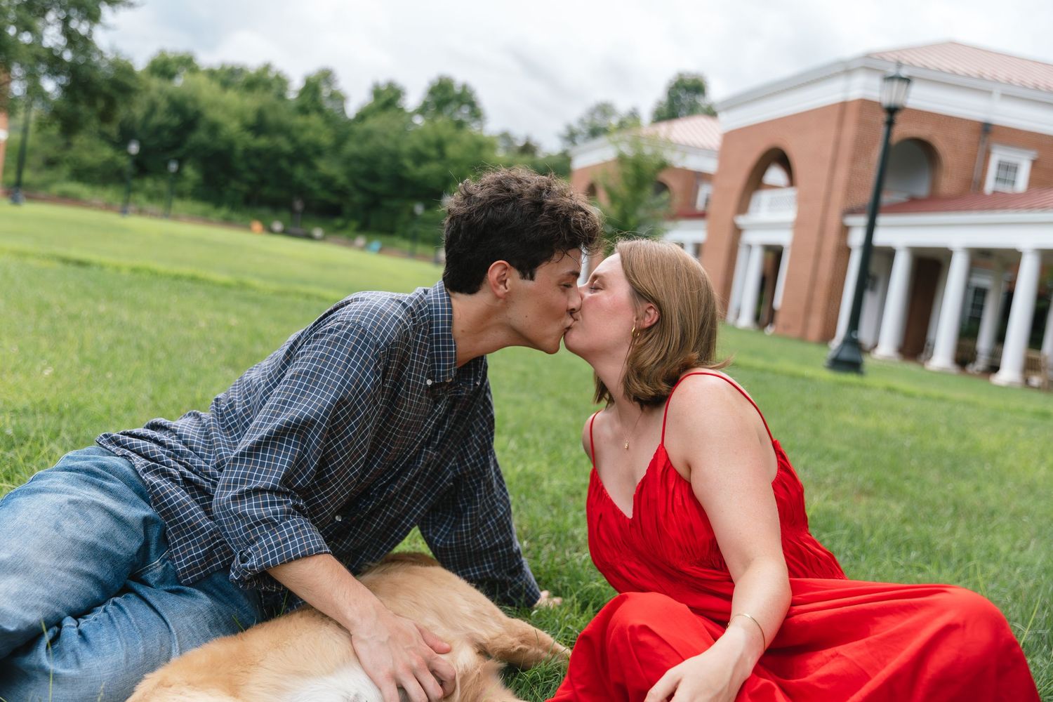 A couple shares a kiss on the grass while their golden dog rests beside them outdoors.