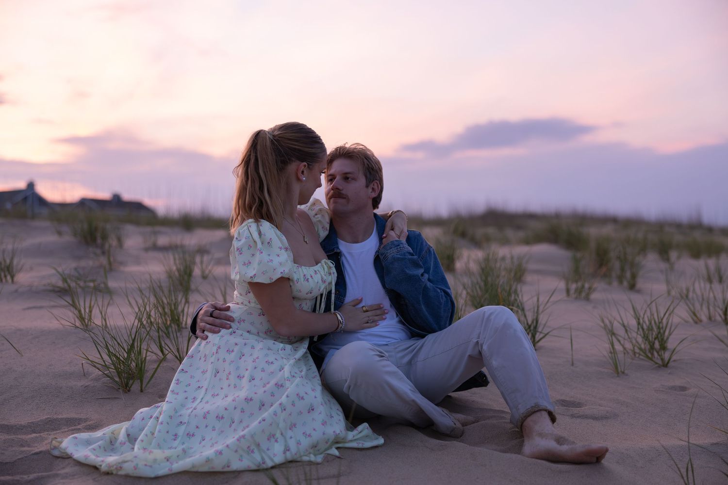Couple leans in for a kiss while sitting on sand dunes during colorful sunset.