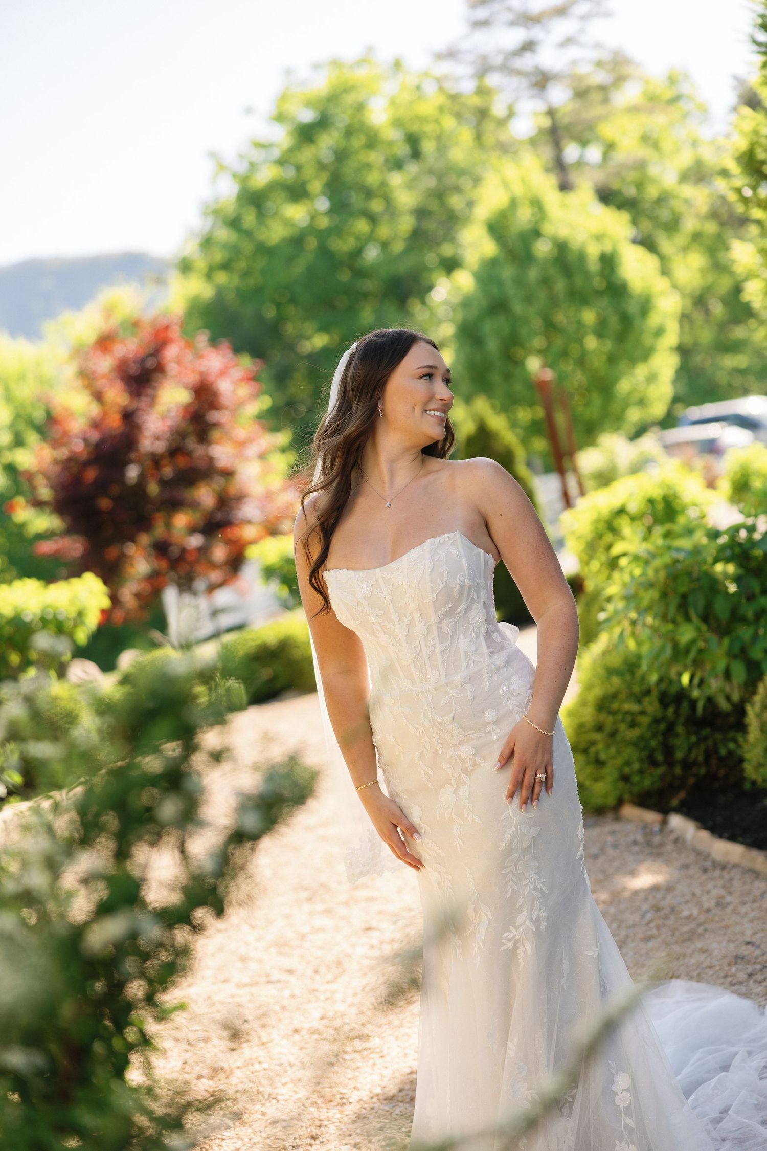 Smiling bride in a strapless lace wedding gown standing in a sunlit garden surrounded by lush greenery.