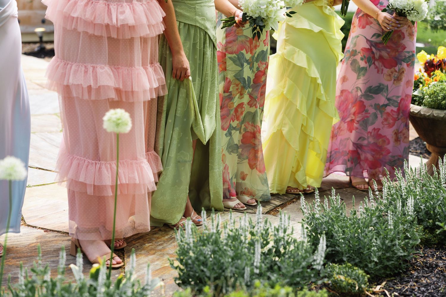 Bridesmaids in colorful mismatched gowns of pink, green, yellow, and floral prints stand in garden.