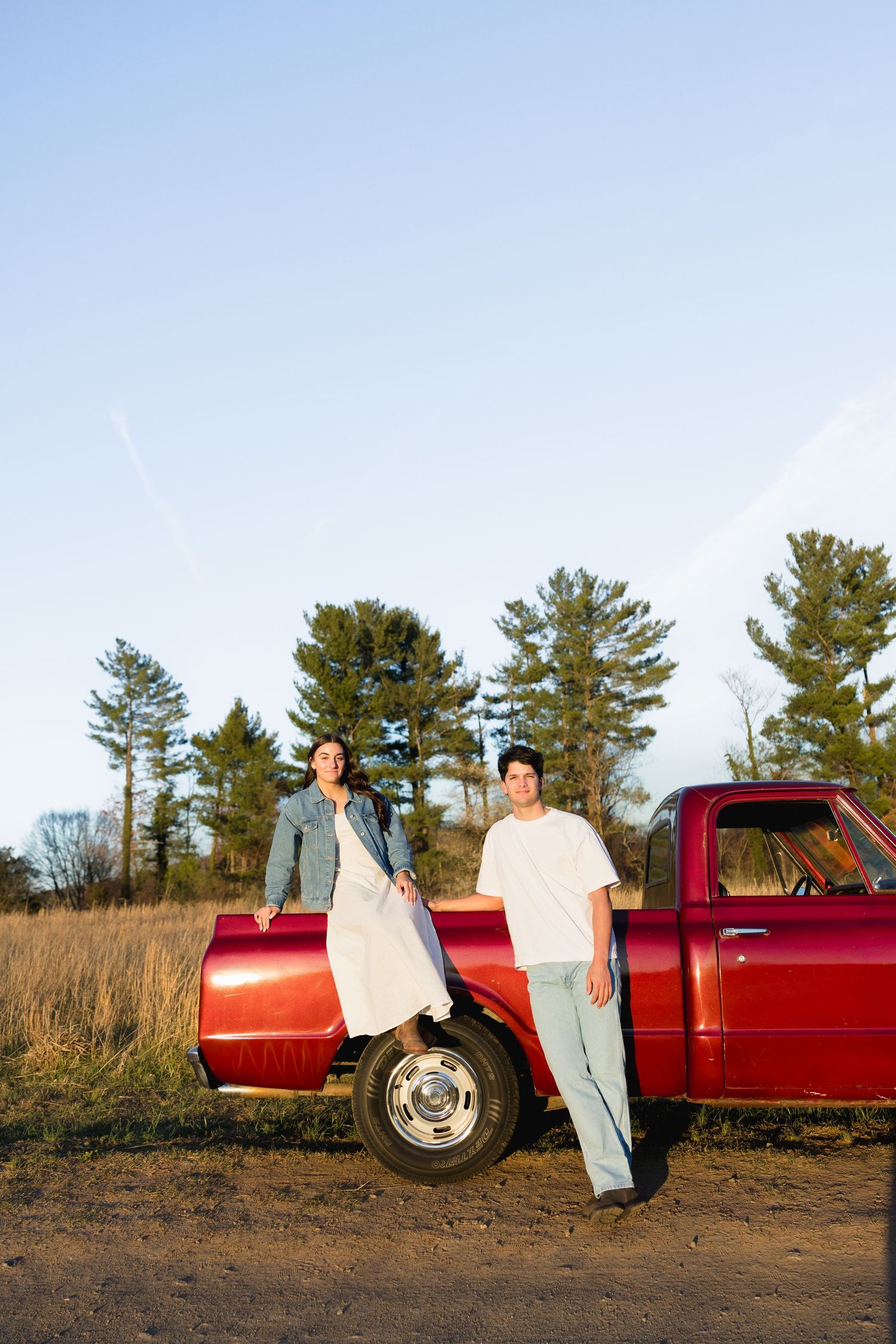 Young couple standing together beside a classic red pickup truck in an open field under a clear blue sky.