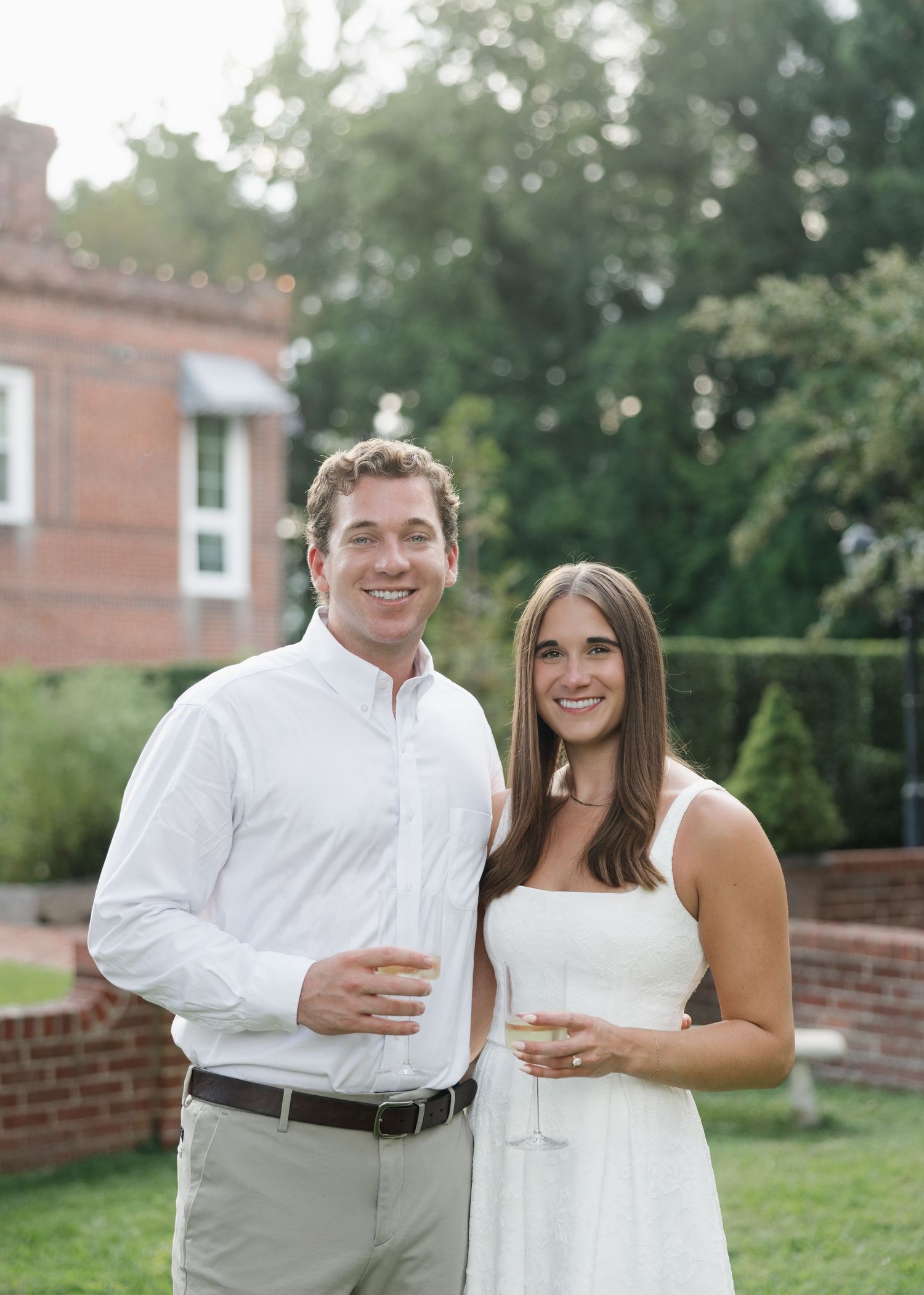 Smiling couple in white attire holding champagne glasses outdoors near a brick building at an evening event.