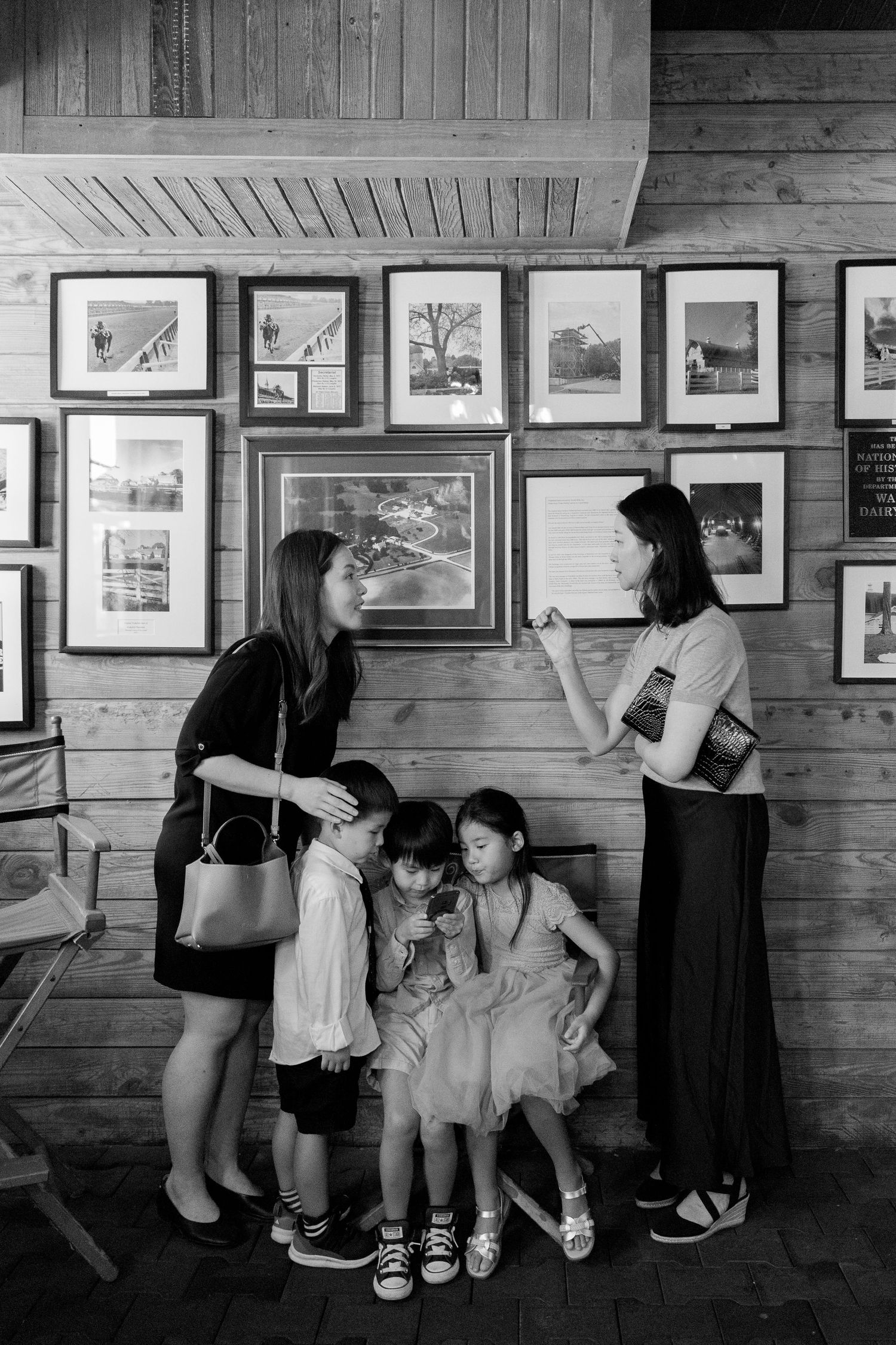 Black and white photo of two women and three children viewing framed artwork on a wooden gallery wall.