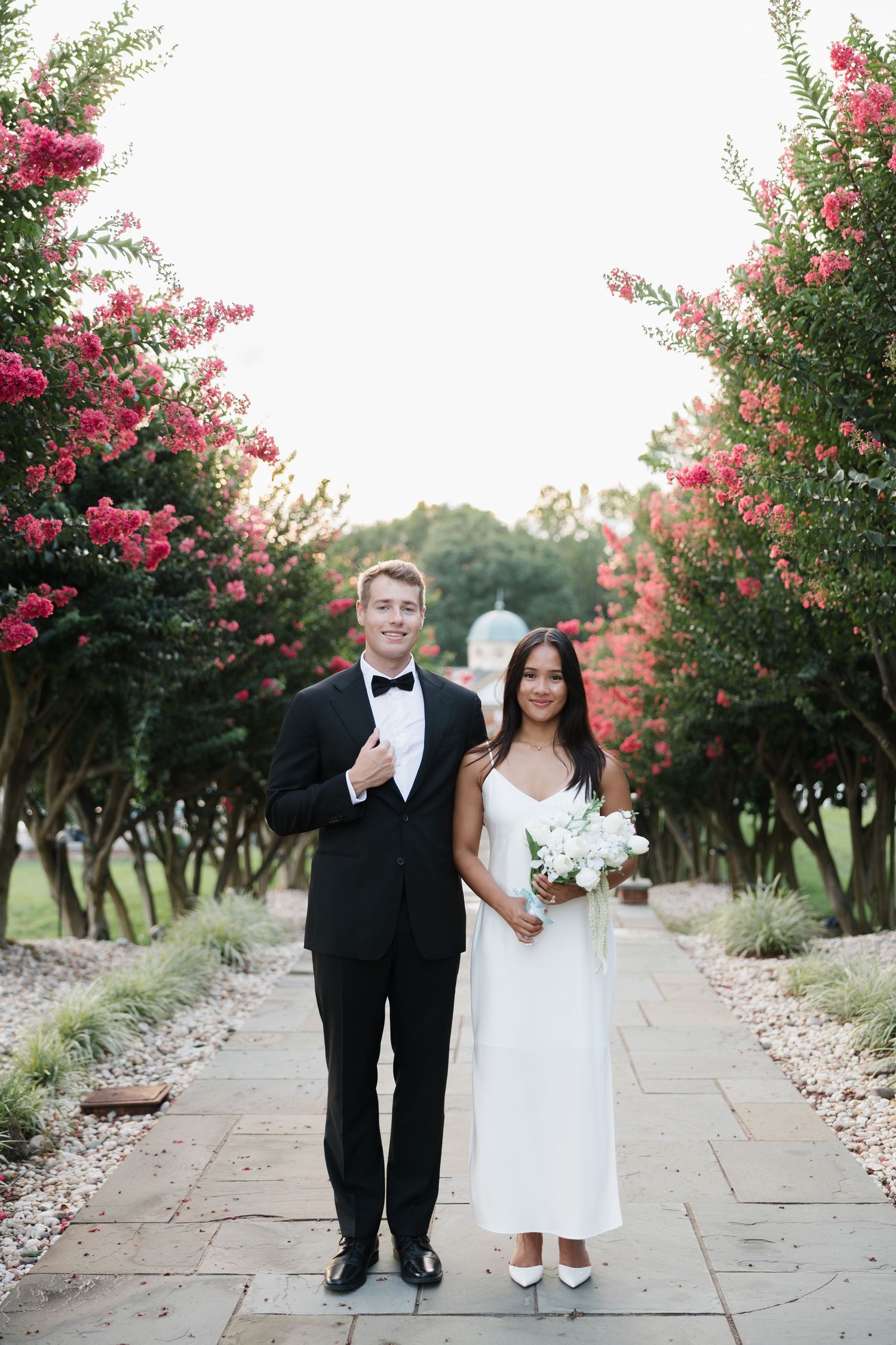 Bride and groom posing on stone path lined with blooming pink crepe myrtle trees on their wedding day.