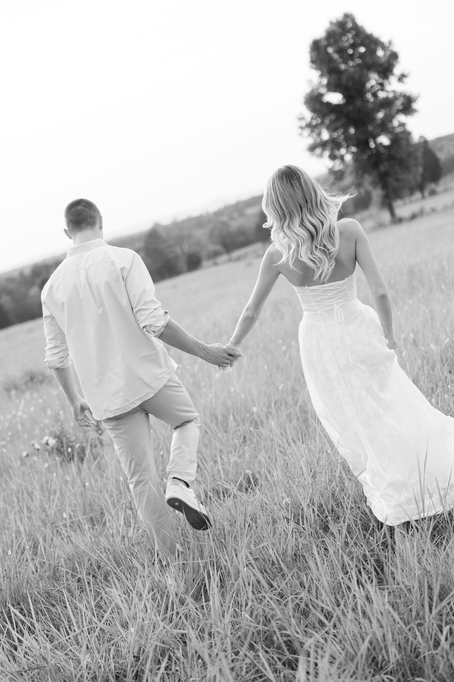 Black and white image of couple holding hands walking through a scenic open field together.