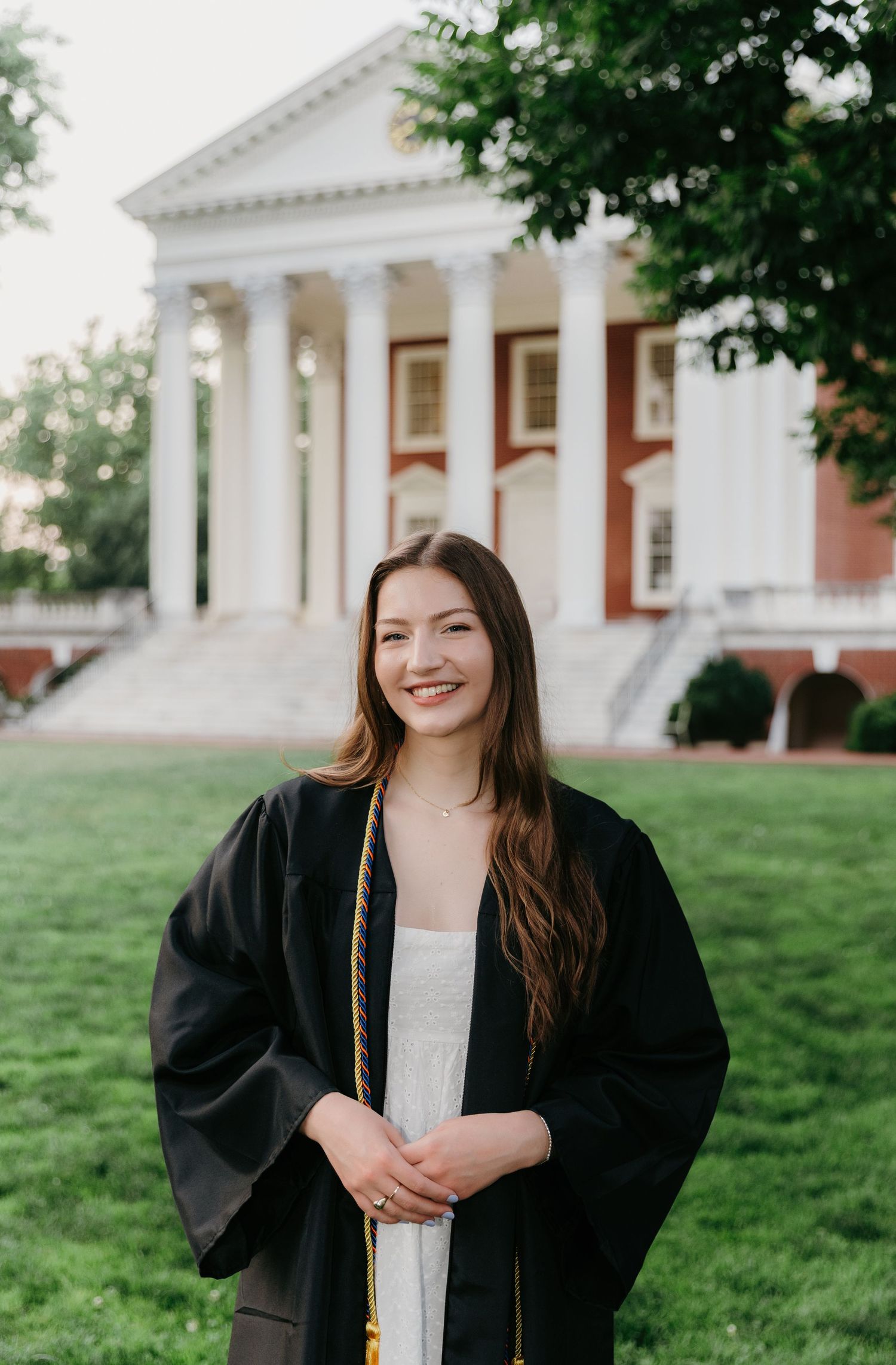 Smiling graduate in black cap and gown stands before a classic columned brick building.