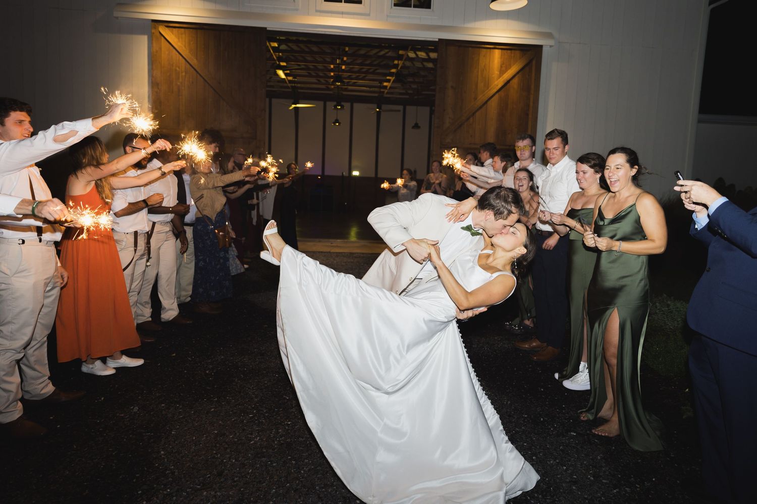 Groom dips bride during sparkler exit at wedding reception surrounded by cheering guests.