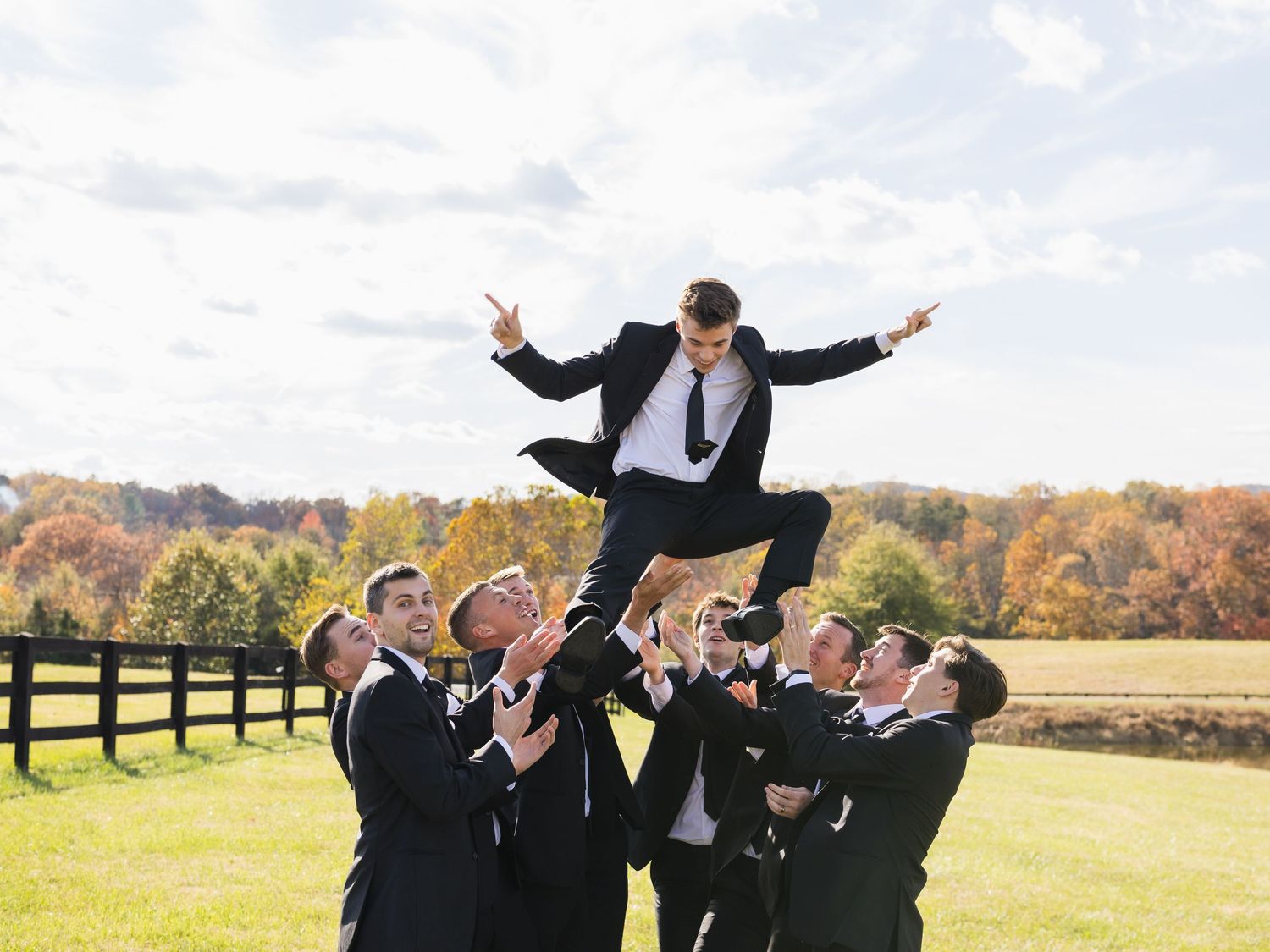 Groomsmen toss a jubilant groom into the air on a sunny autumn day at an outdoor wedding venue.