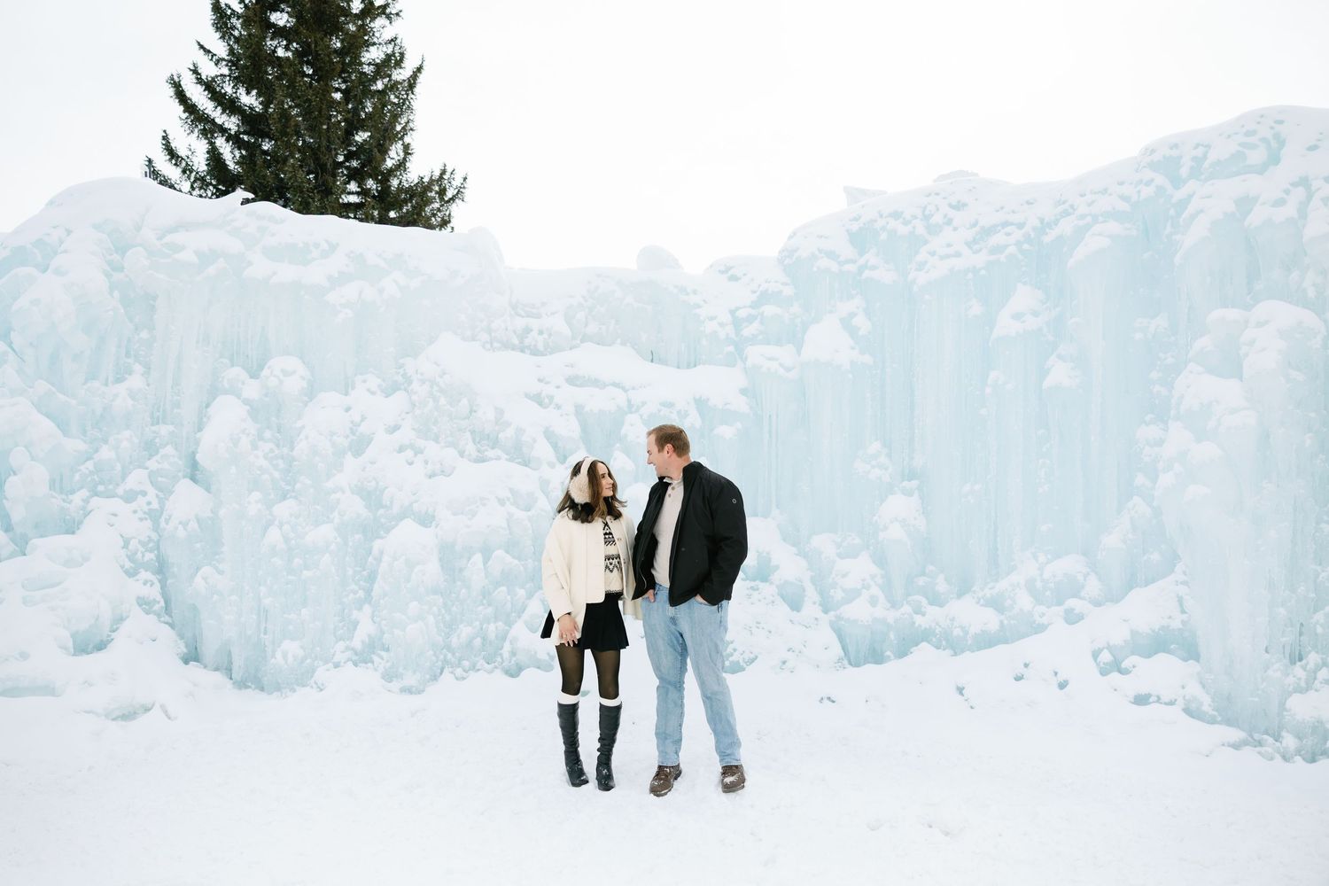 Couple holding hands while standing in front of a large frozen ice castle and snow-covered pine tree.