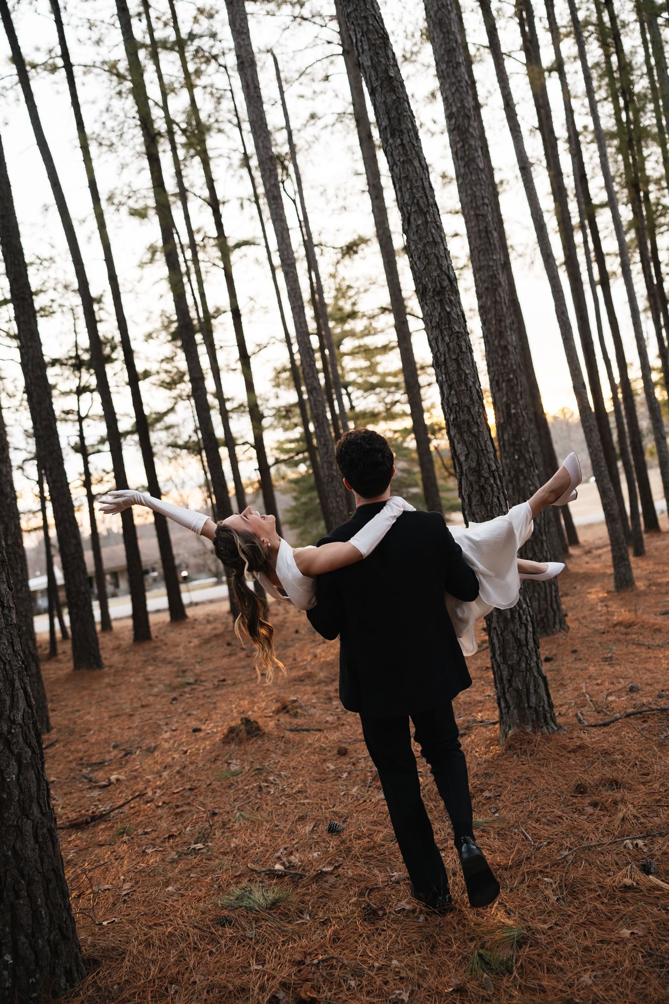 Groom carries bride with arms outstretched through sunlit pine forest in joyful wedding portrait.