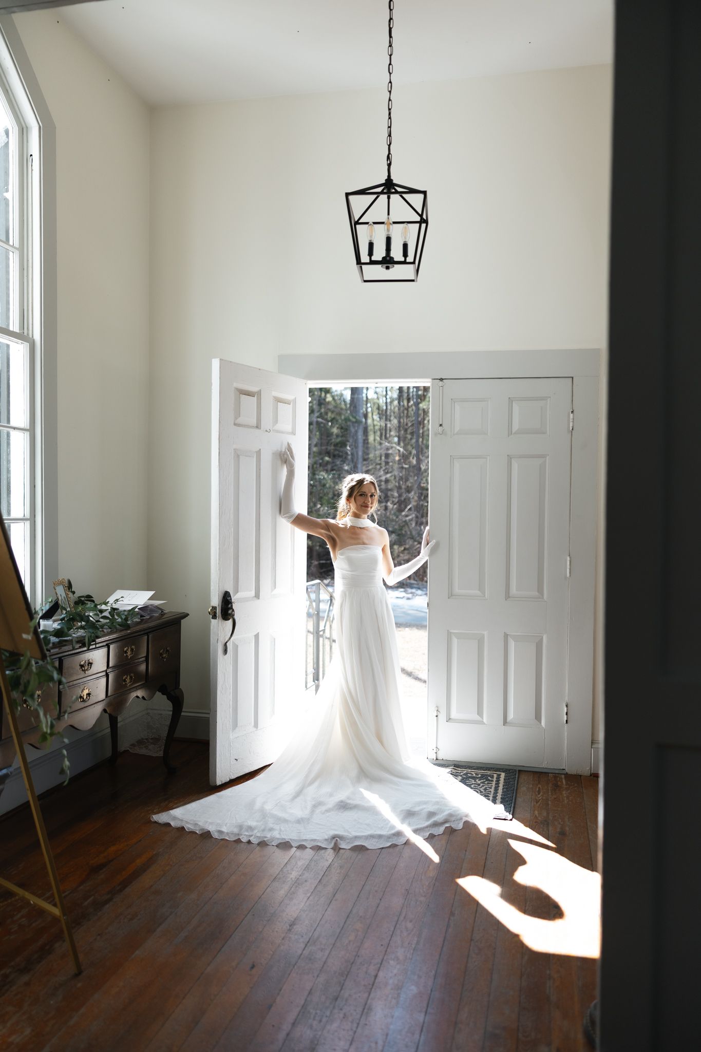 Bride in white wedding gown with long train standing in sunlit doorway of elegant home.
