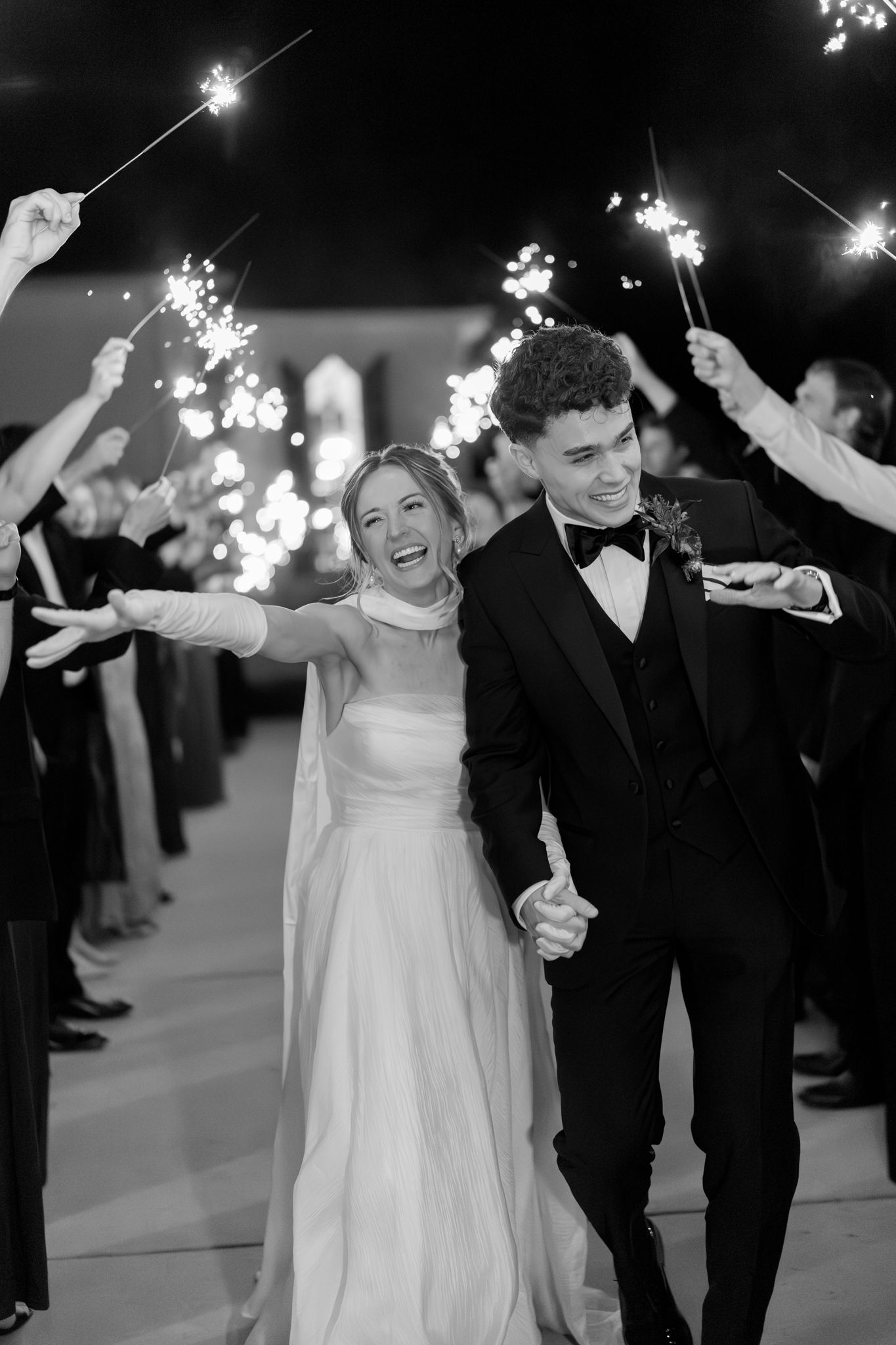 Black and white photo of laughing bride and groom walking through sparkler send-off with guests cheering around them.