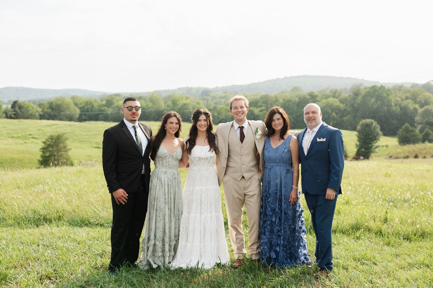 Wedding party of six posing in lush green meadow with rolling hills, bride in white gown, guests in formal attire.