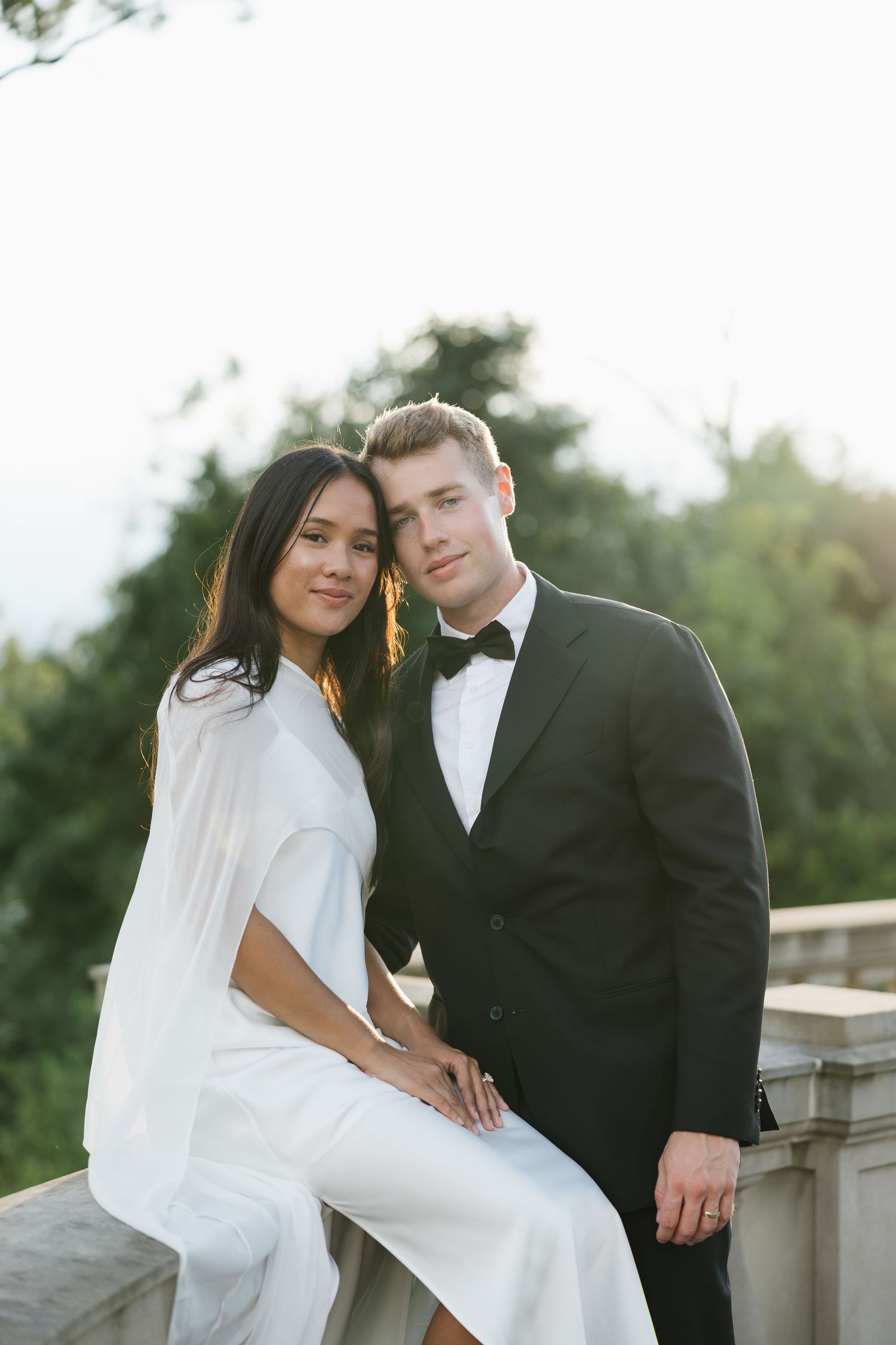 A couple in wedding attire poses outdoors, the bride in white and groom in a black tuxedo, against a lush green backdrop.