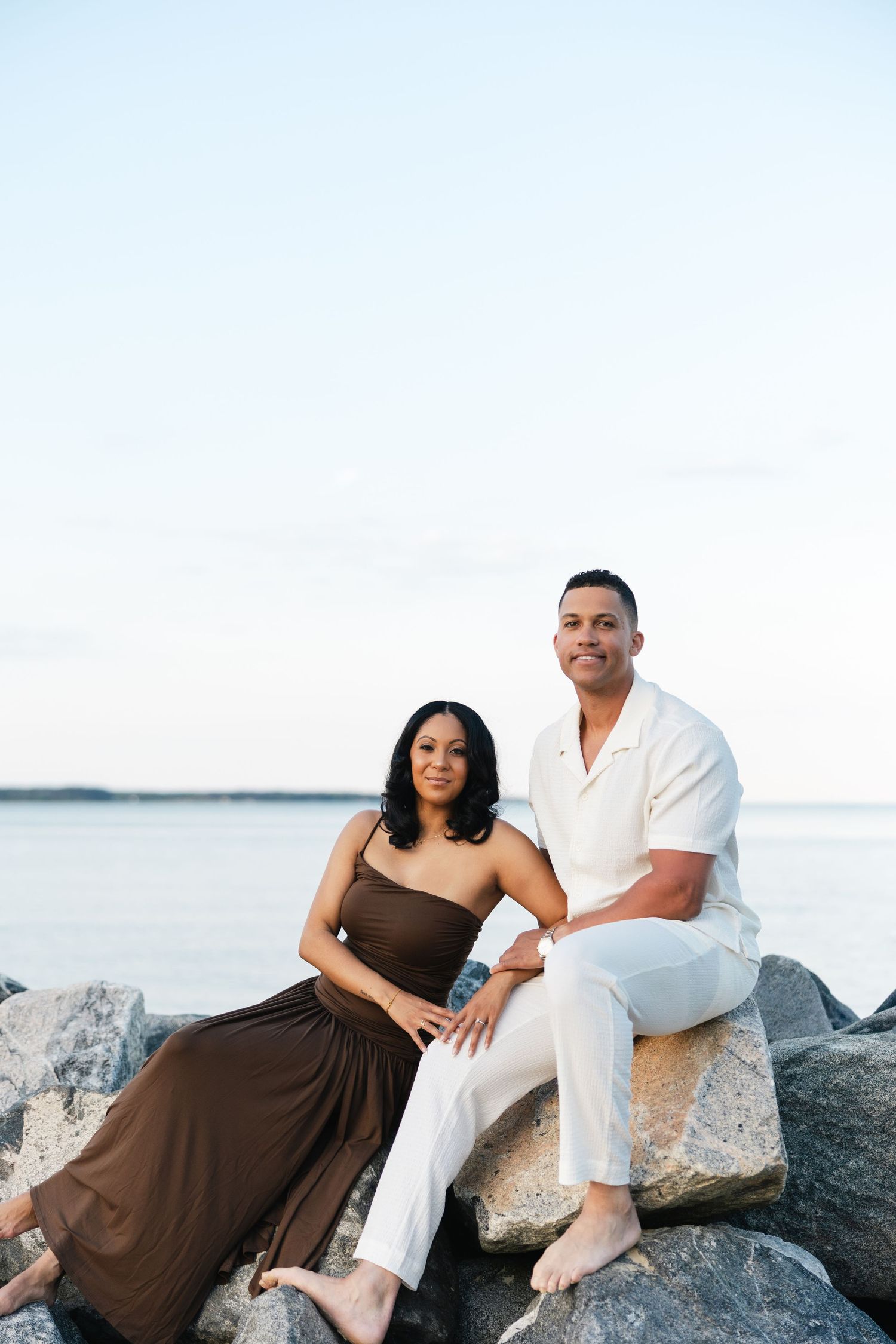 Happy couple sitting on rocks by the ocean, woman in brown dress, man in white outfit.