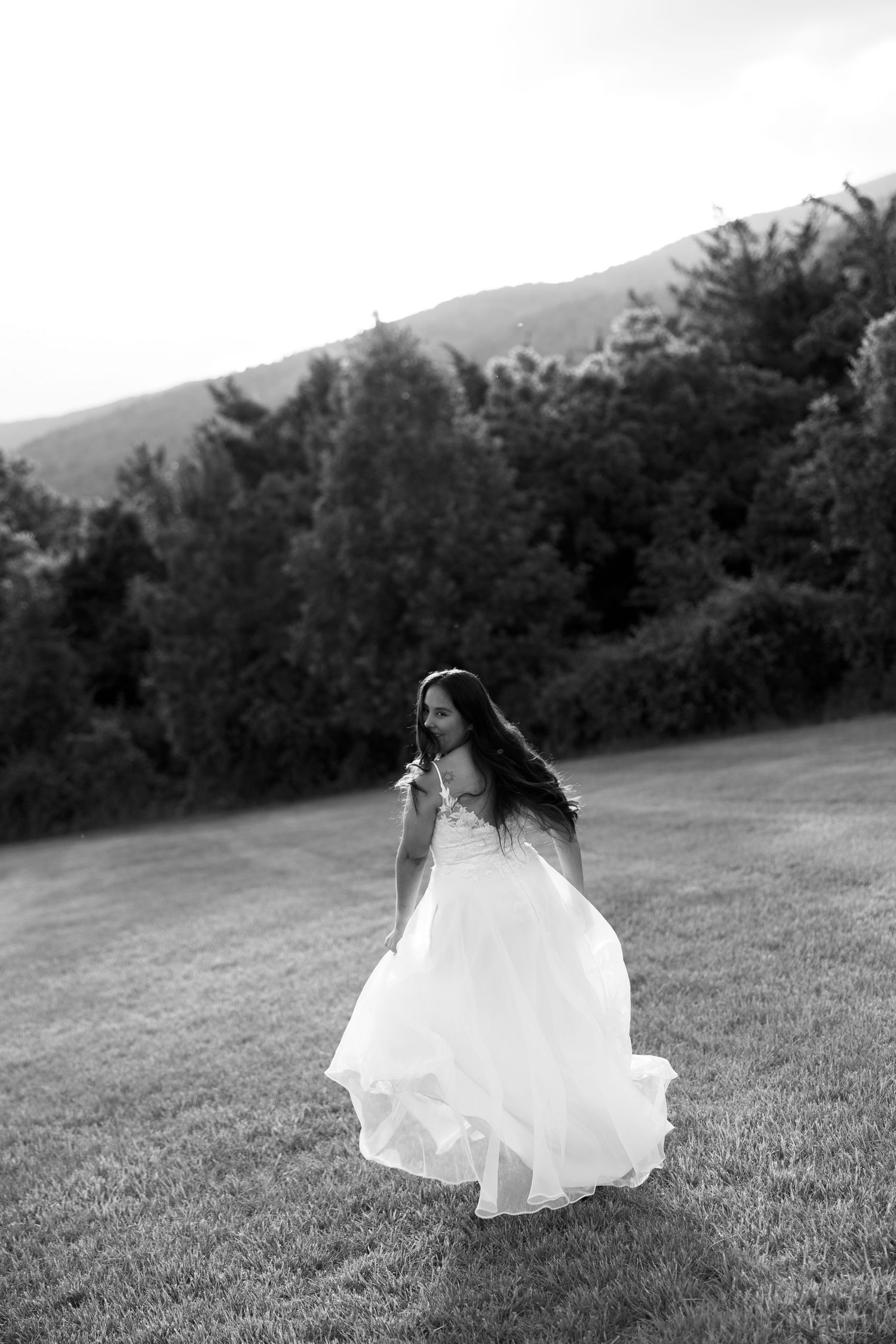 Black and white photo of a bride in a flowing white gown twirling on a green lawn with mountains in the background.