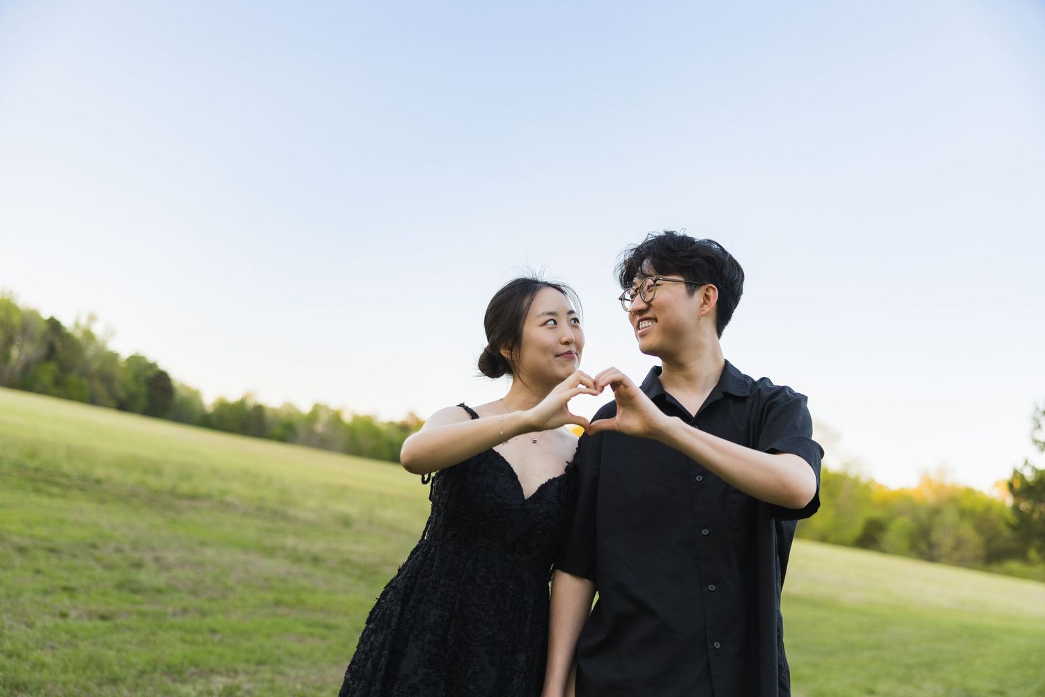A couple makes a heart shape with their hands while smiling at each other in a green park field.