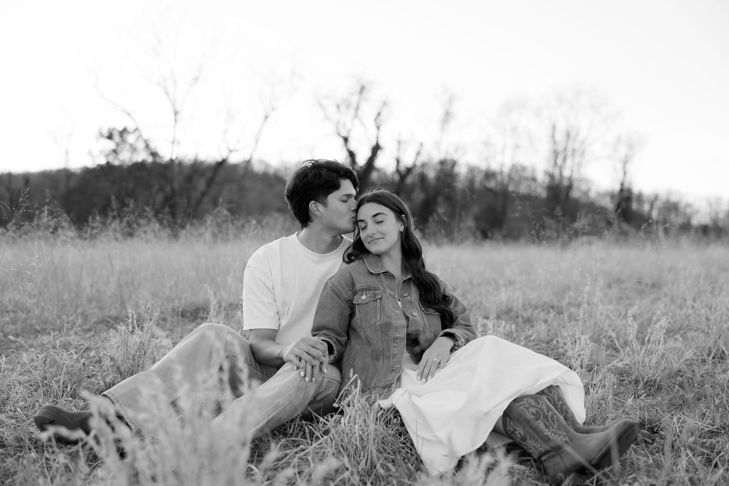A black and white photo of a couple sitting in a field, the man kissing the womans cheek at sunset.