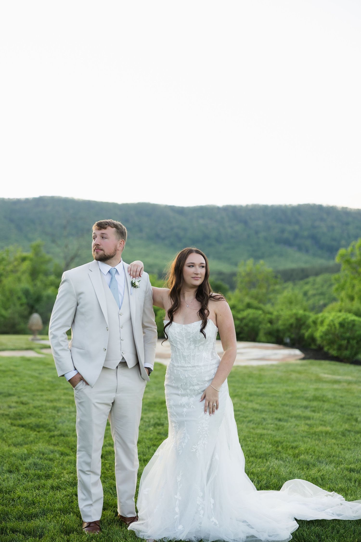 Bride and groom pose outdoors in white wedding attire with lush green mountain backdrop.