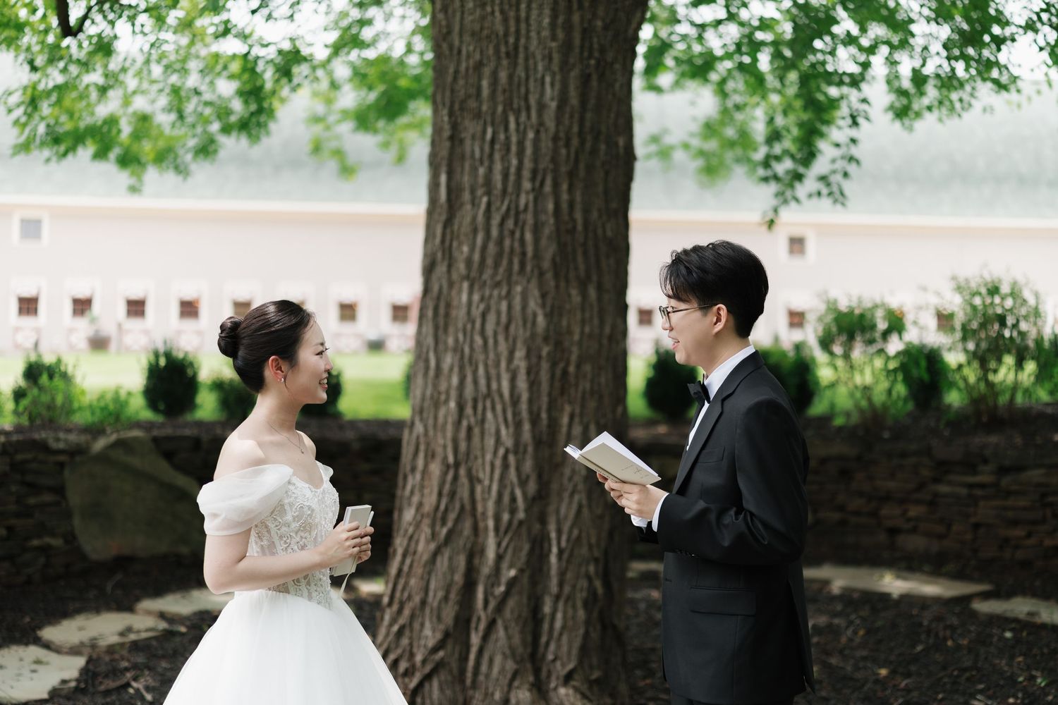 Bride in white gown and groom in black tuxedo exchange vows under a large tree in a lush garden setting.