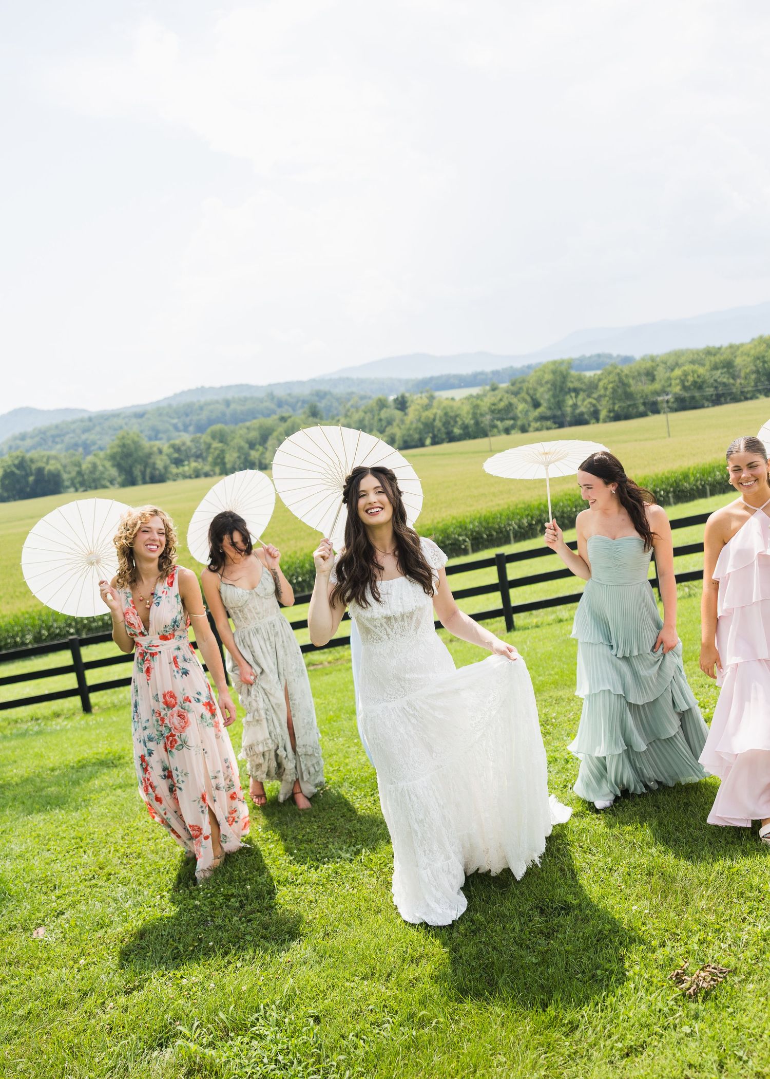 Bride and bridesmaids in colorful dresses holding white parasols walking on green grass outdoors.