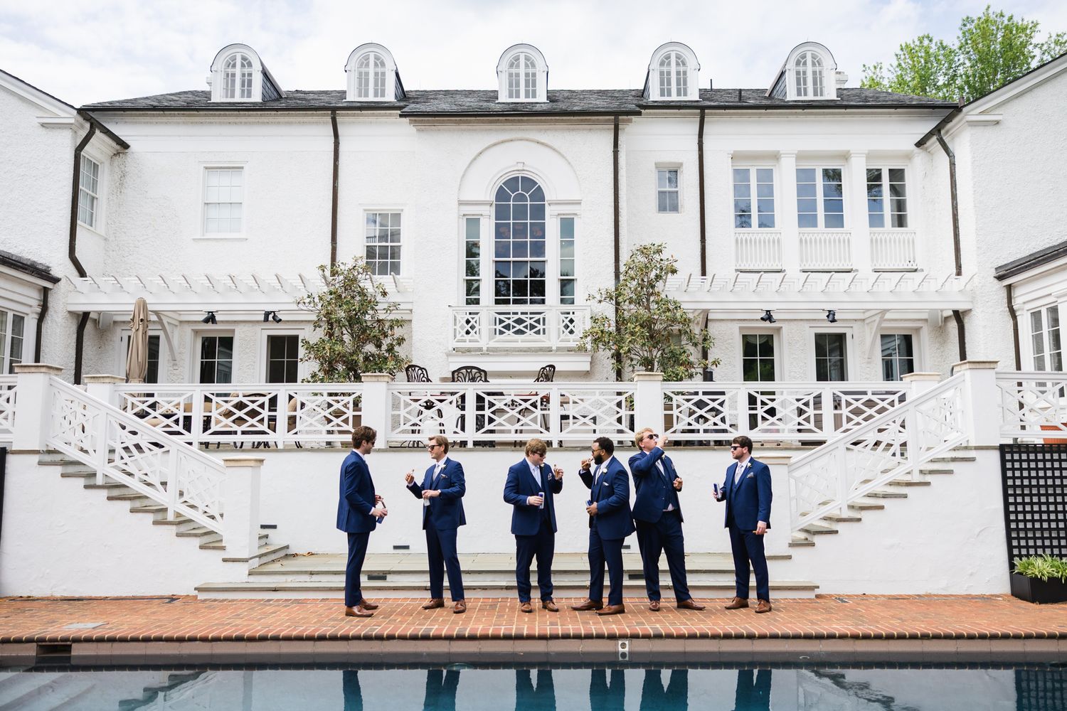 Groomsmen in navy suits gather and chat beside a pool in front of an elegant white mansion on a wedding day.