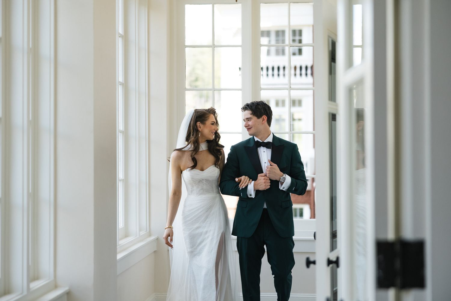 Bride in white gown and groom in dark tuxedo walking together through elegant white hallway.
