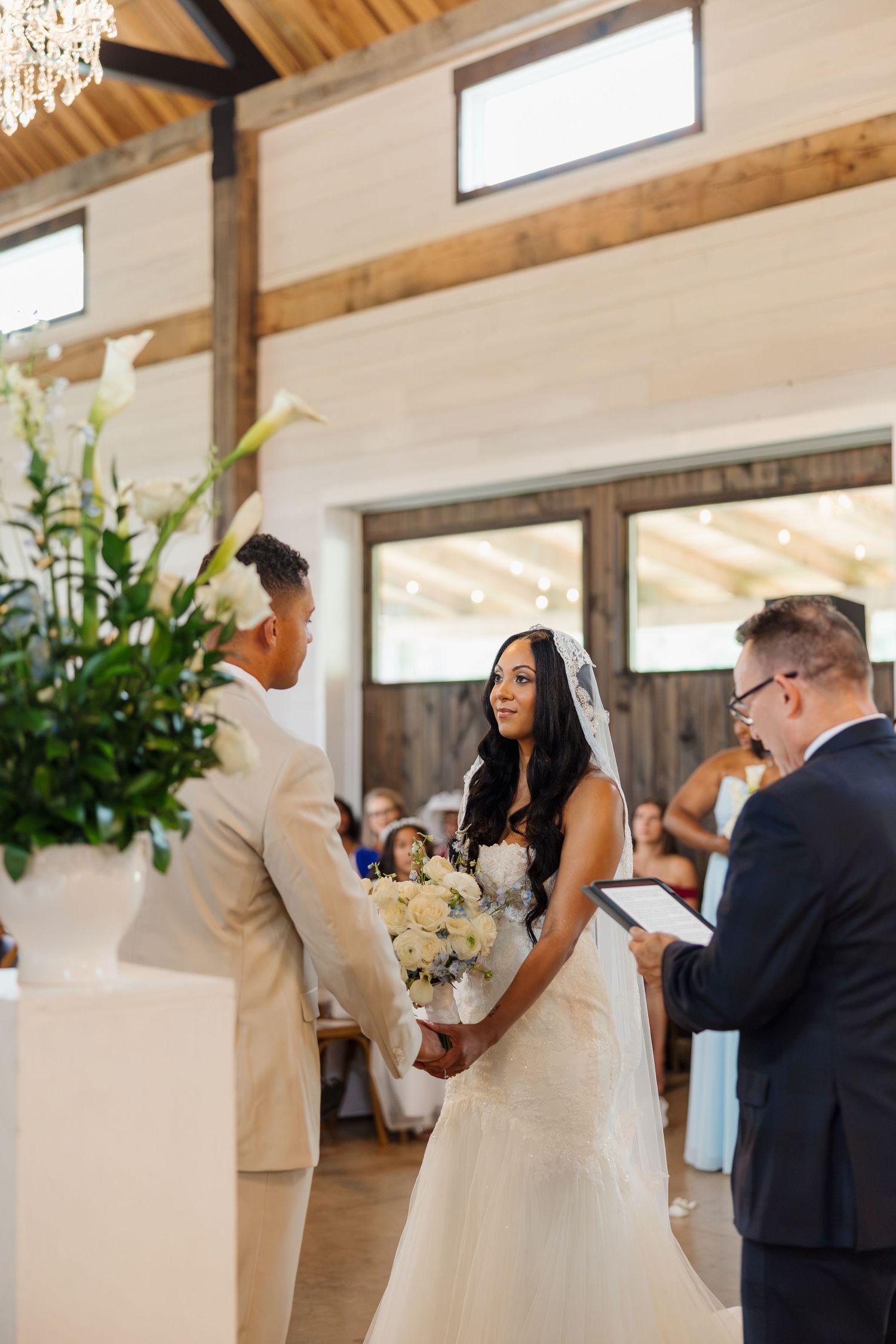 Bride in white gown holding bouquet exchanges vows with groom in beige suit at elegant barn wedding ceremony.