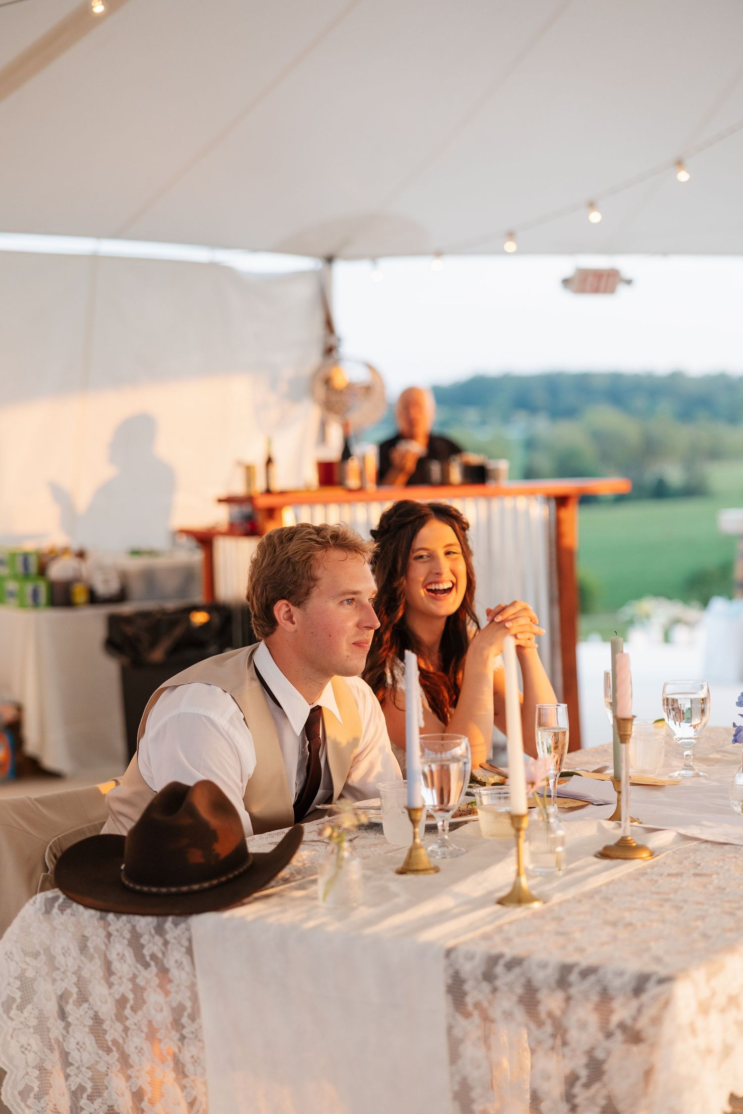 Bride and groom laugh joyfully at their wedding reception table under a white tent at golden hour.
