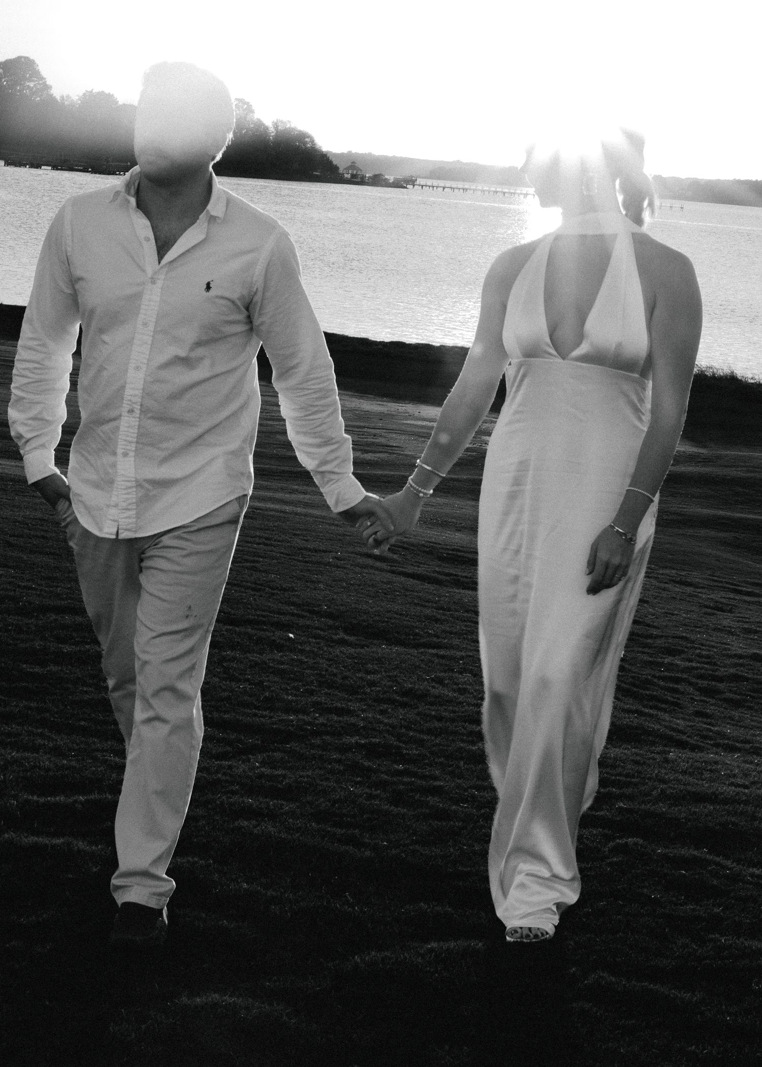 Black and white photo of a couple holding hands walking on a beach at sunset, wearing white outfits.