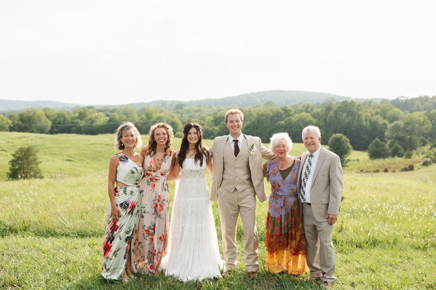 Wedding party poses in a lush green field, bride in white gown with groom and family members in floral dresses.
