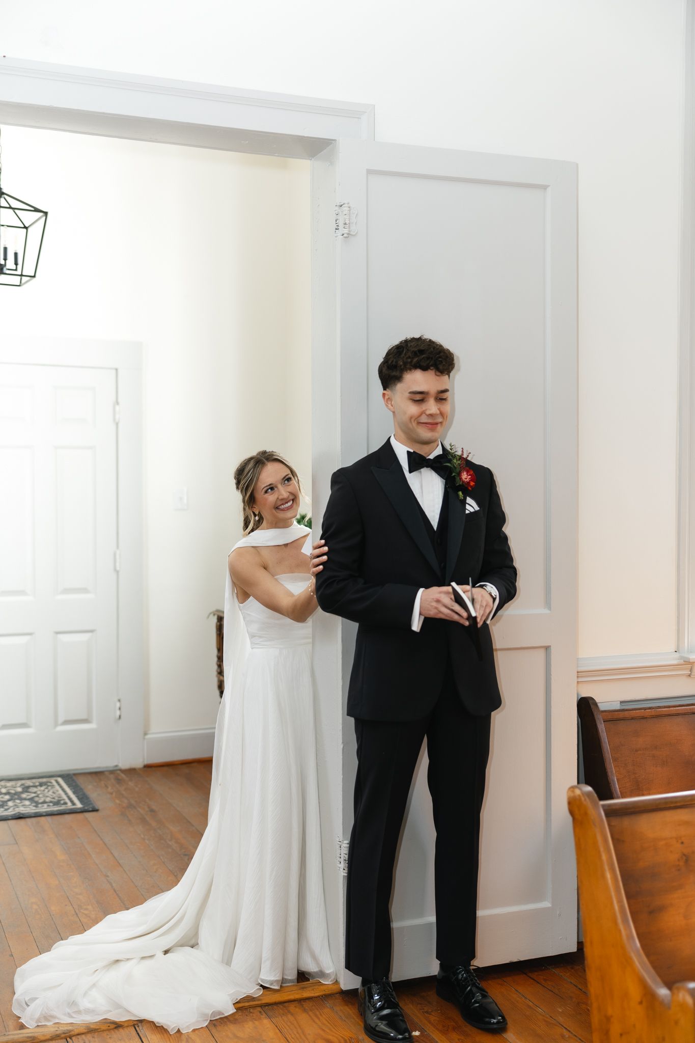 Bride in white gown sharing first look moment with groom in black tuxedo indoors.