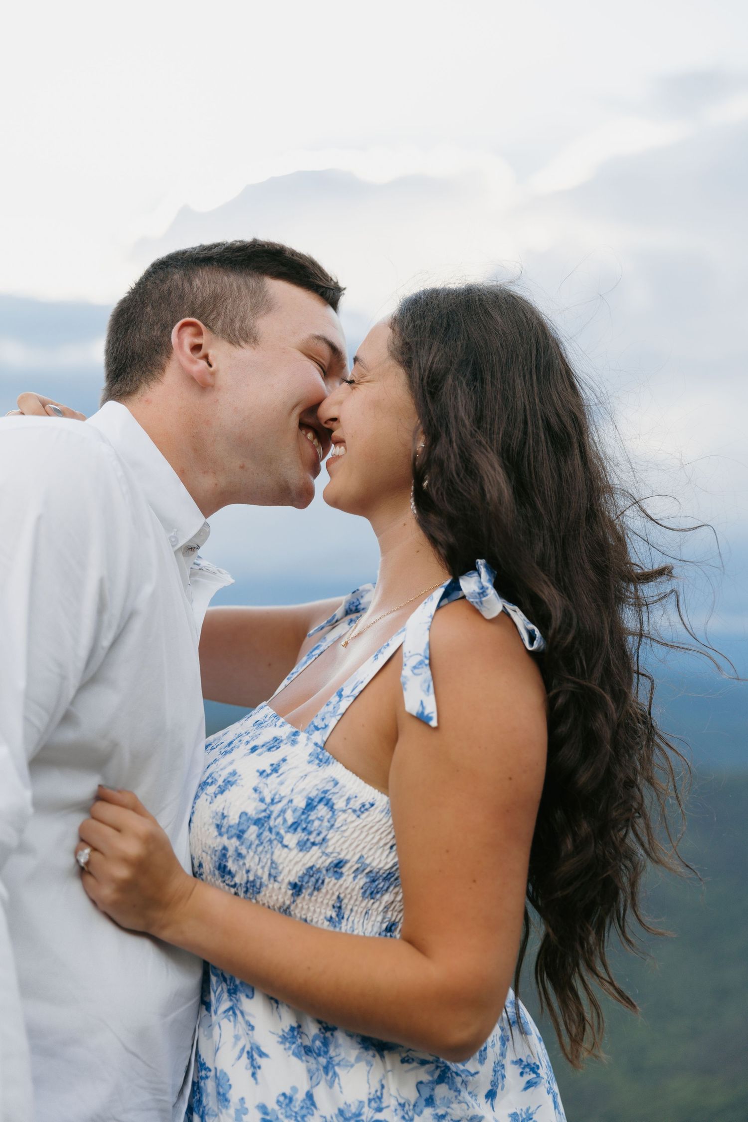 A joyful couple shares a kiss with mountains and cloudy skies in the background during an outdoor photo session.